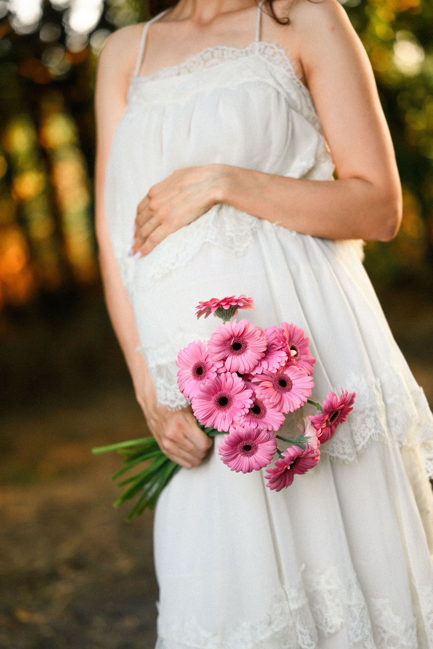 Pregnant woman in white lace dress holding pink gerbera daisy bouquet outdoors in soft natural light.