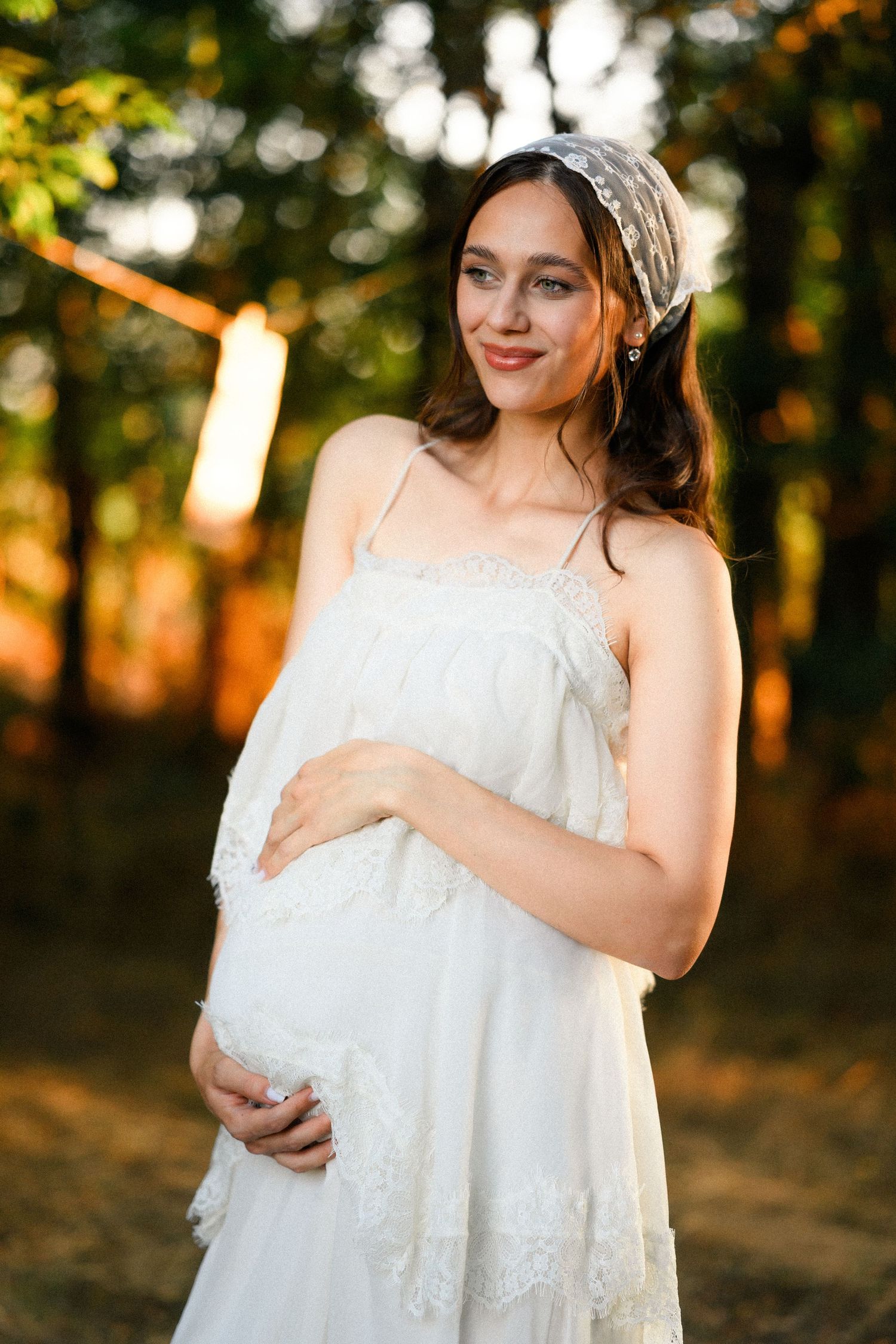 Smiling pregnant woman in white lace dress holding baby bump outdoors in golden sunset forest light.
