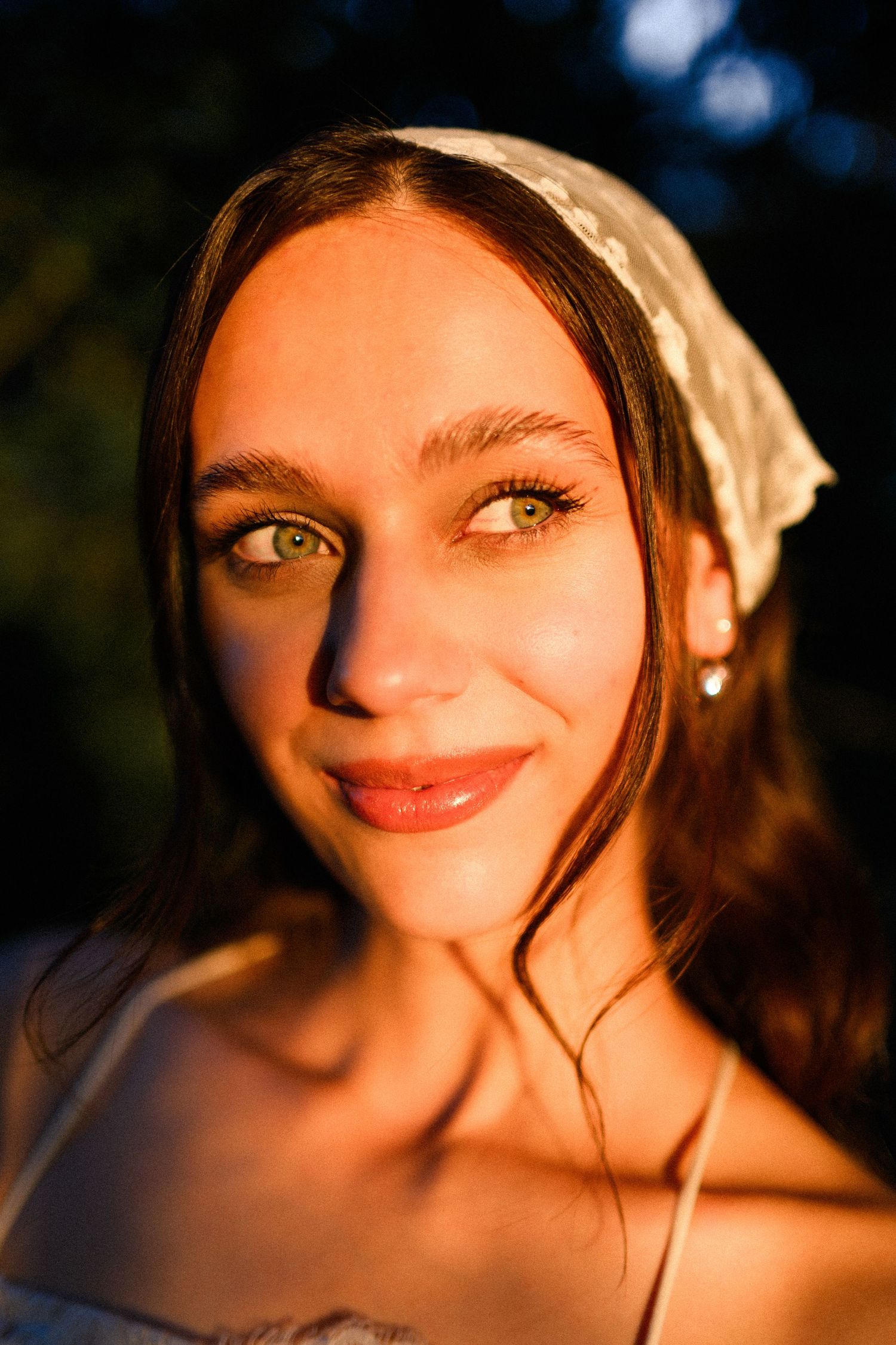 Young woman with green eyes and white headscarf bathed in warm golden hour sunlight, smiling softly outdoors.