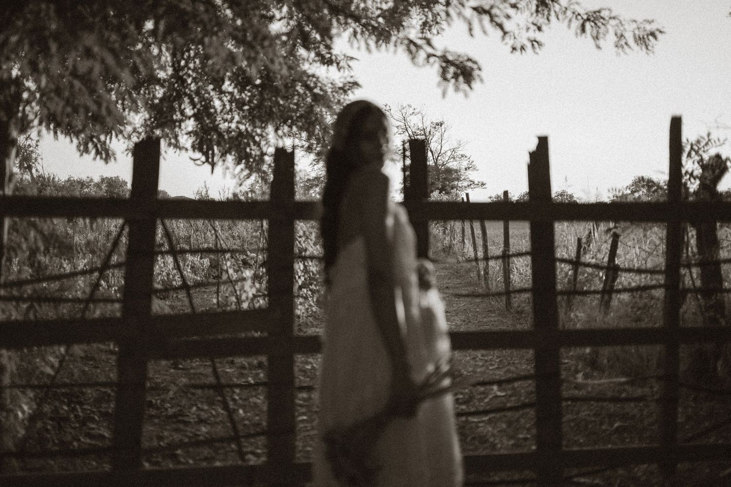 A woman in a white dress stands near a rustic wooden fence in a moody, sepia-toned outdoor portrait.