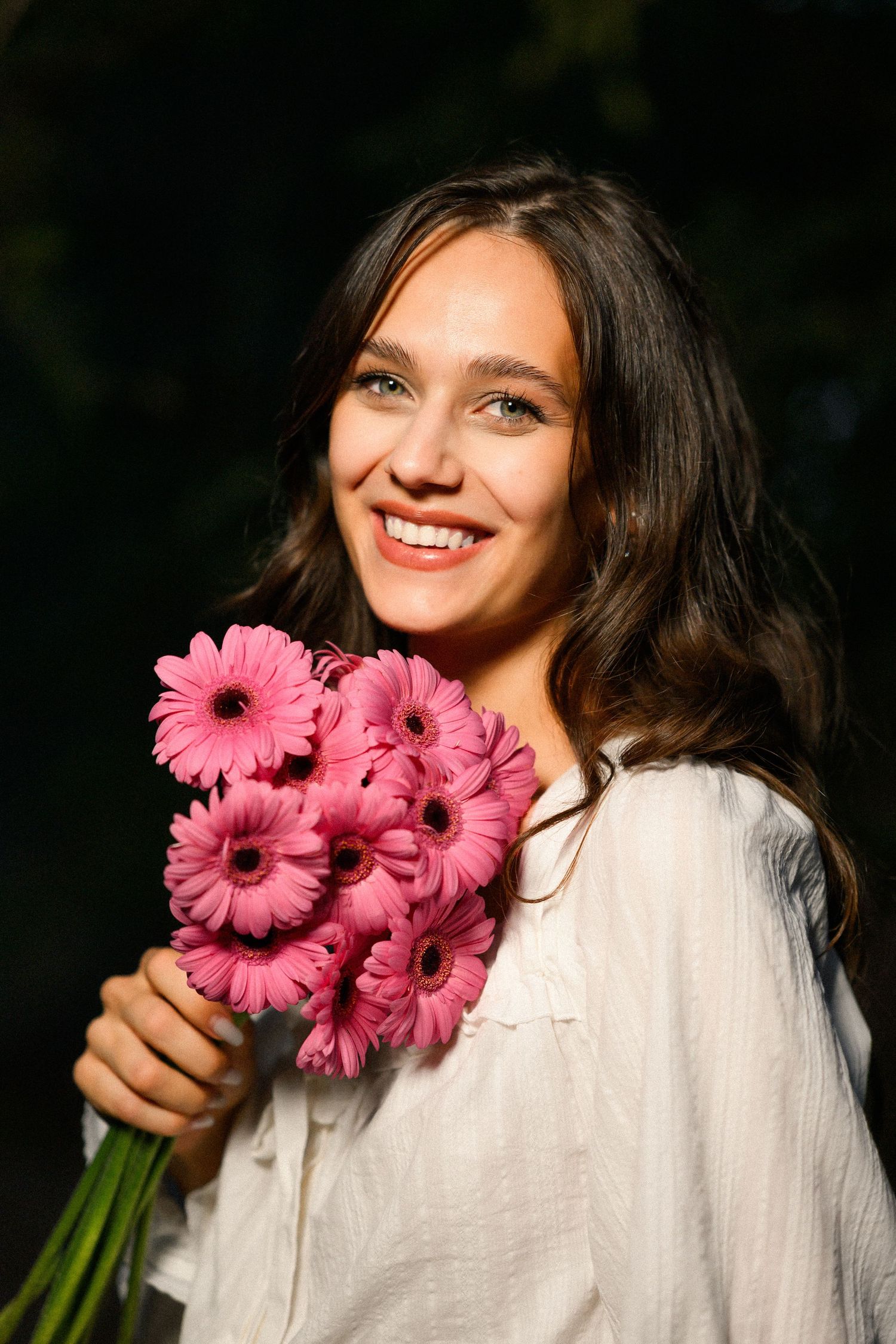 Smiling young woman holding a bouquet of pink gerbera daisies outdoors in natural sunlight.