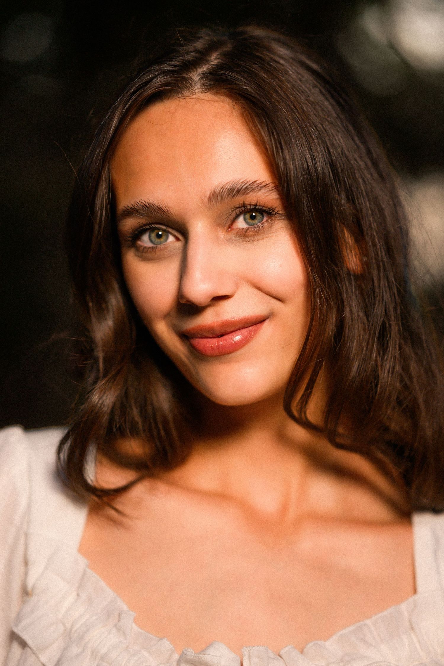Young woman with dark hair and green eyes smiling softly outdoors in warm golden sunlight.