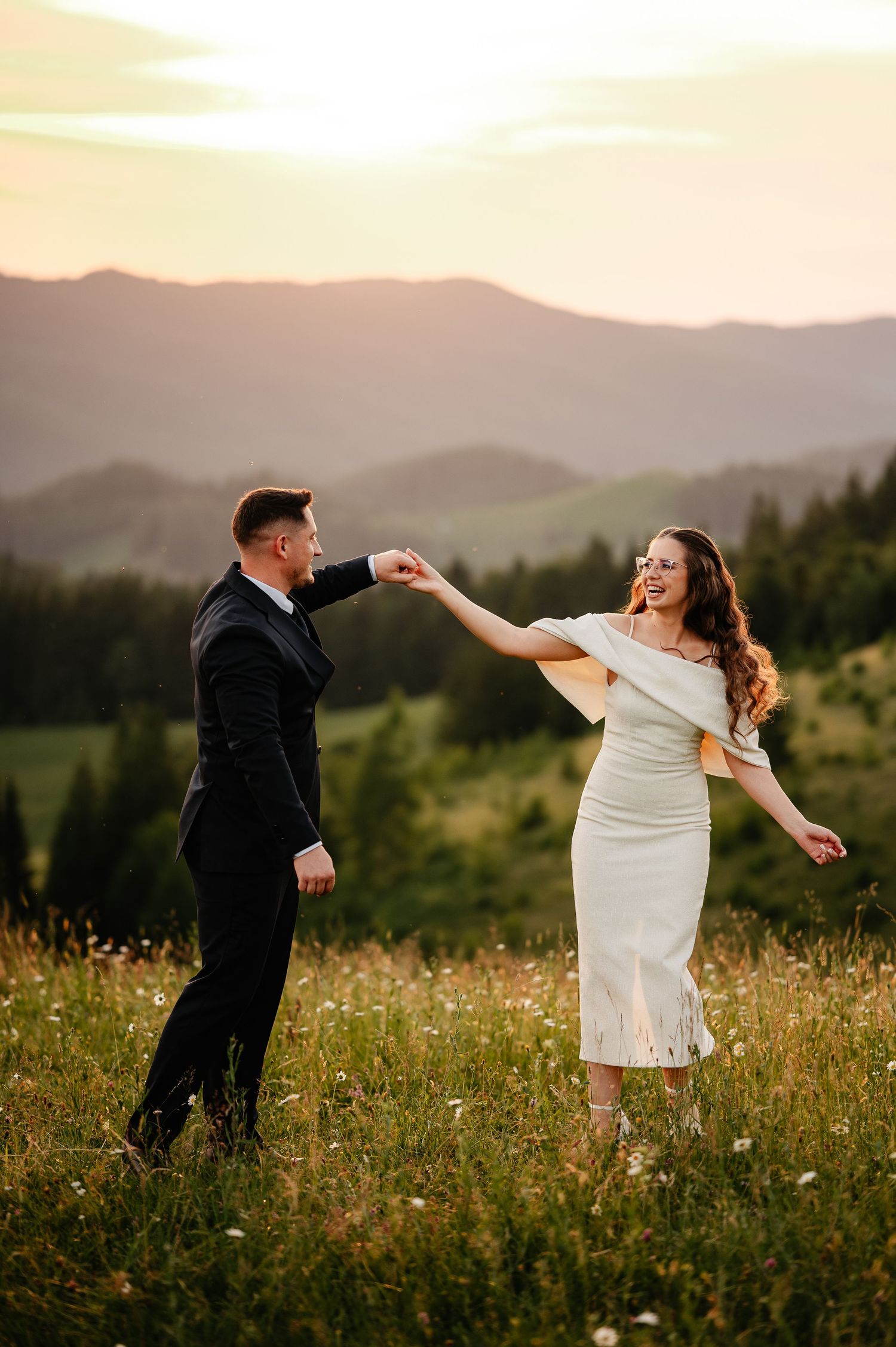 Couple dancing in a meadow at sunset with mountain views, man in suit and woman in white midi dress.