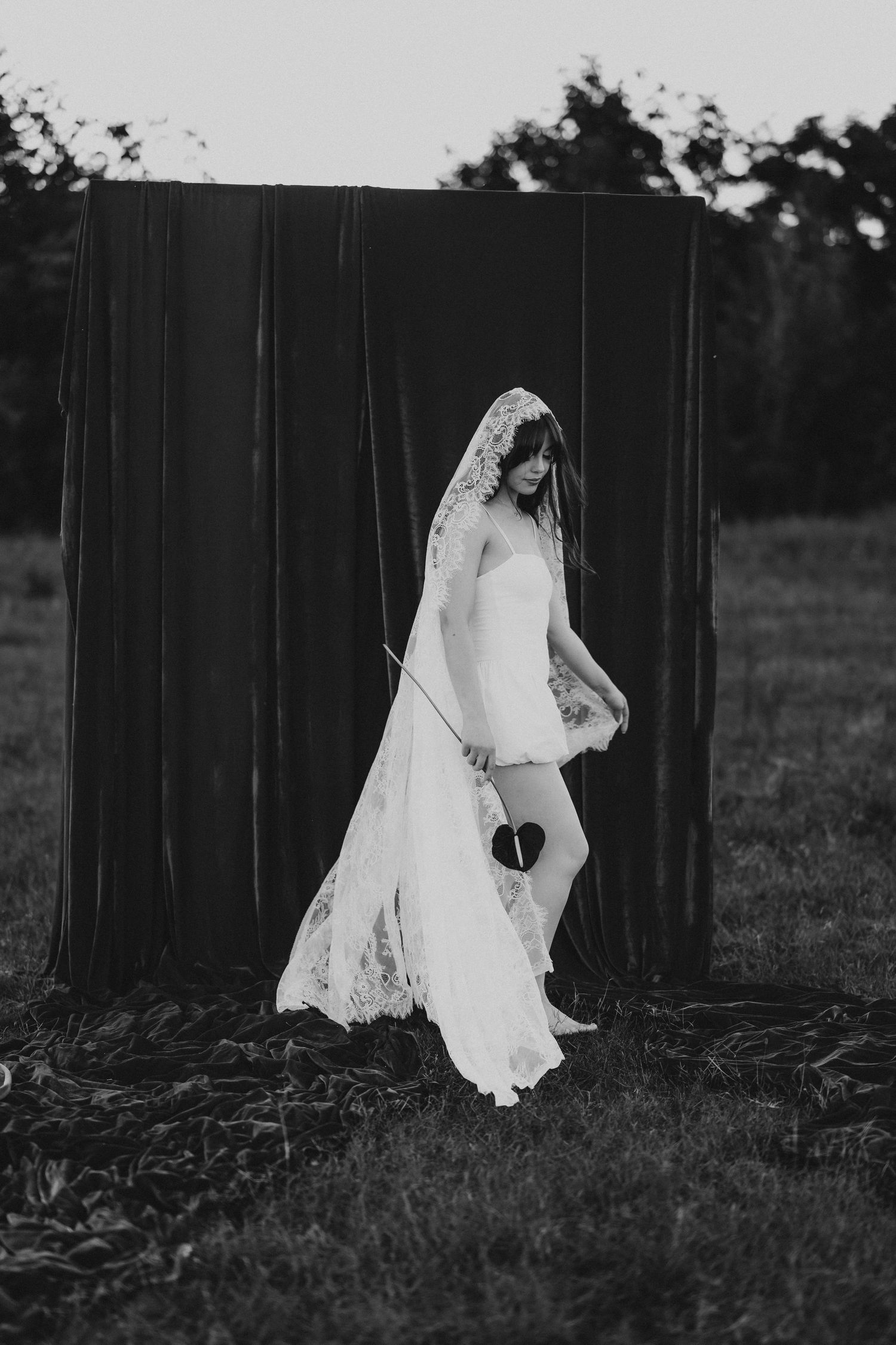 Black and white photo of a bride in lace veil and white dress standing before dark fabric backdrop outdoors.