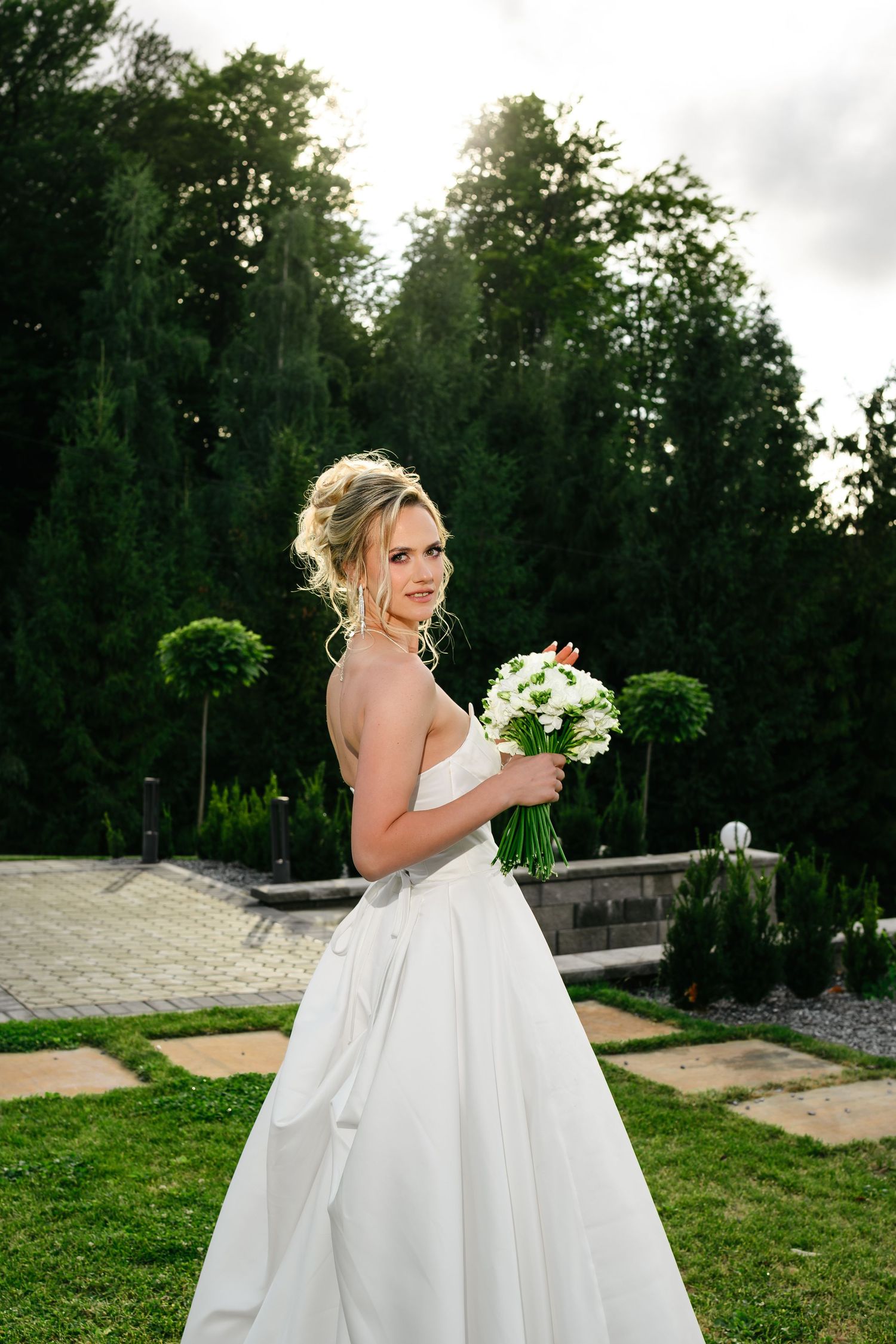 Smiling bride in white strapless ball gown holding white flower bouquet in lush green garden setting.