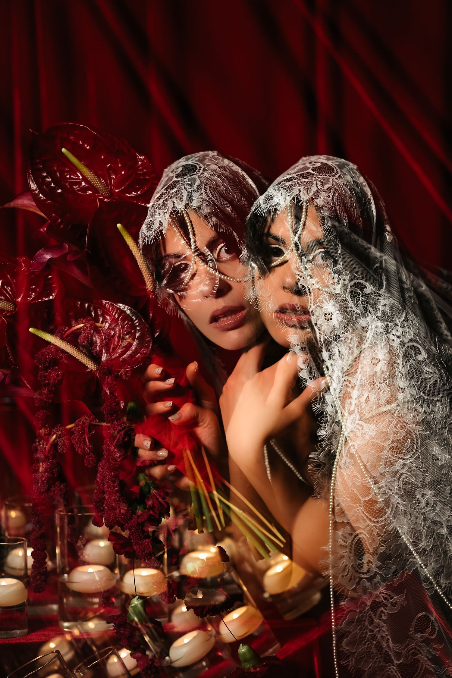 Two women wearing ornate white lace veils pose among red anthurium flowers and candles against crimson velvet draping.