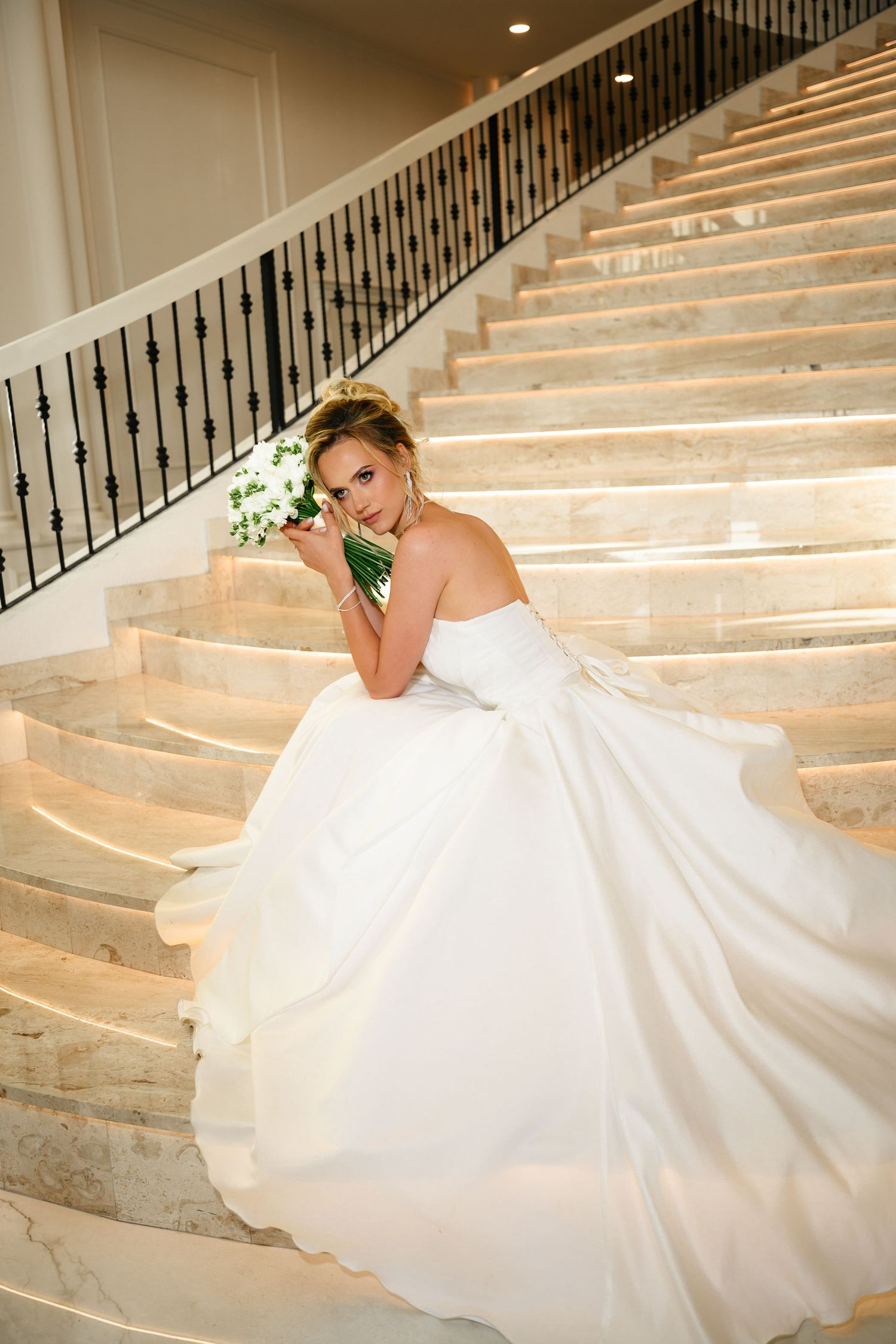 Bride in a flowing white ball gown sitting on marble stairs, holding a white bouquet, looking elegantly over her shoulder.