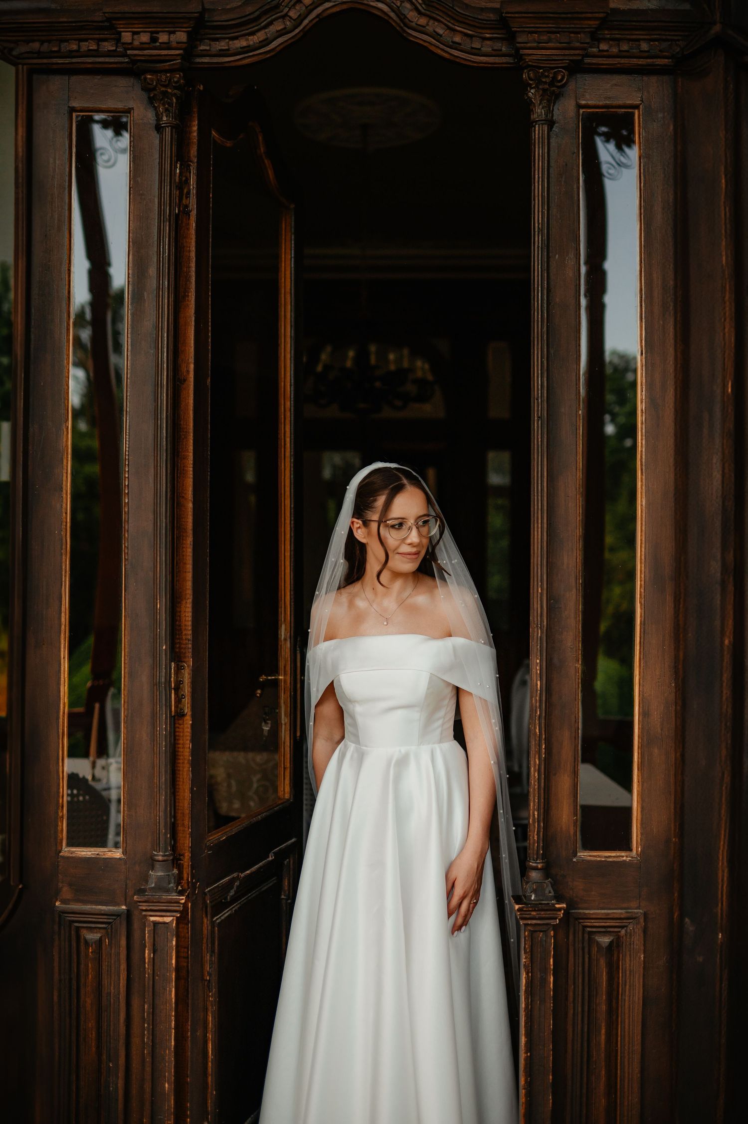 Bride in off-shoulder white gown and veil poses between carved wooden doors at golden hour.