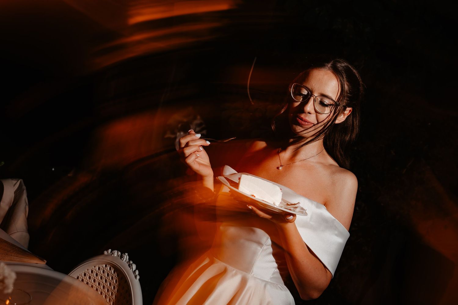 Bride in elegant white off-shoulder gown enjoying wedding cake with dreamy swirling light effects at night.