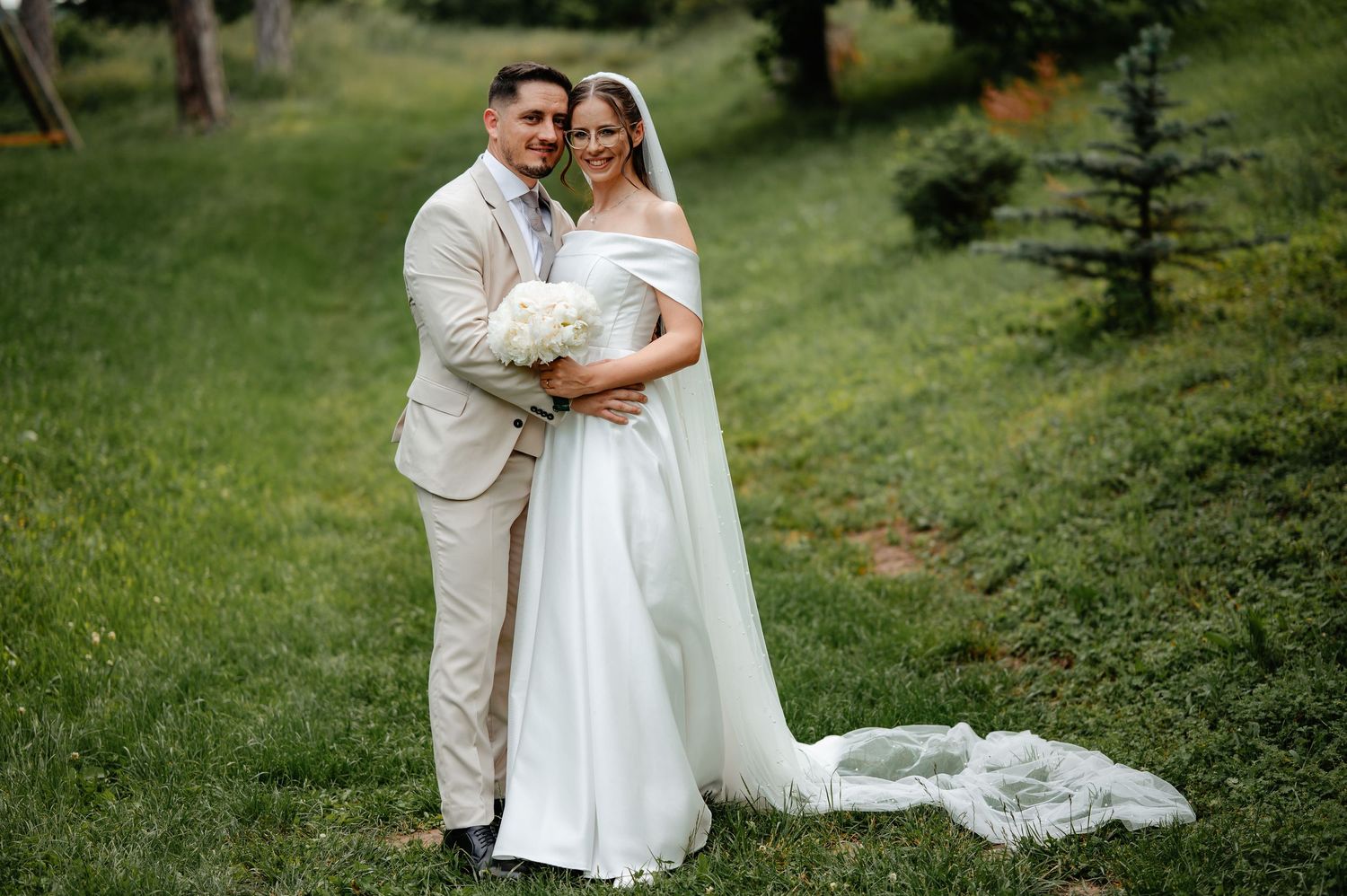 Bride and groom pose together on green grass, she holds white bouquet in elegant wedding gown with veil, he wears beige suit.