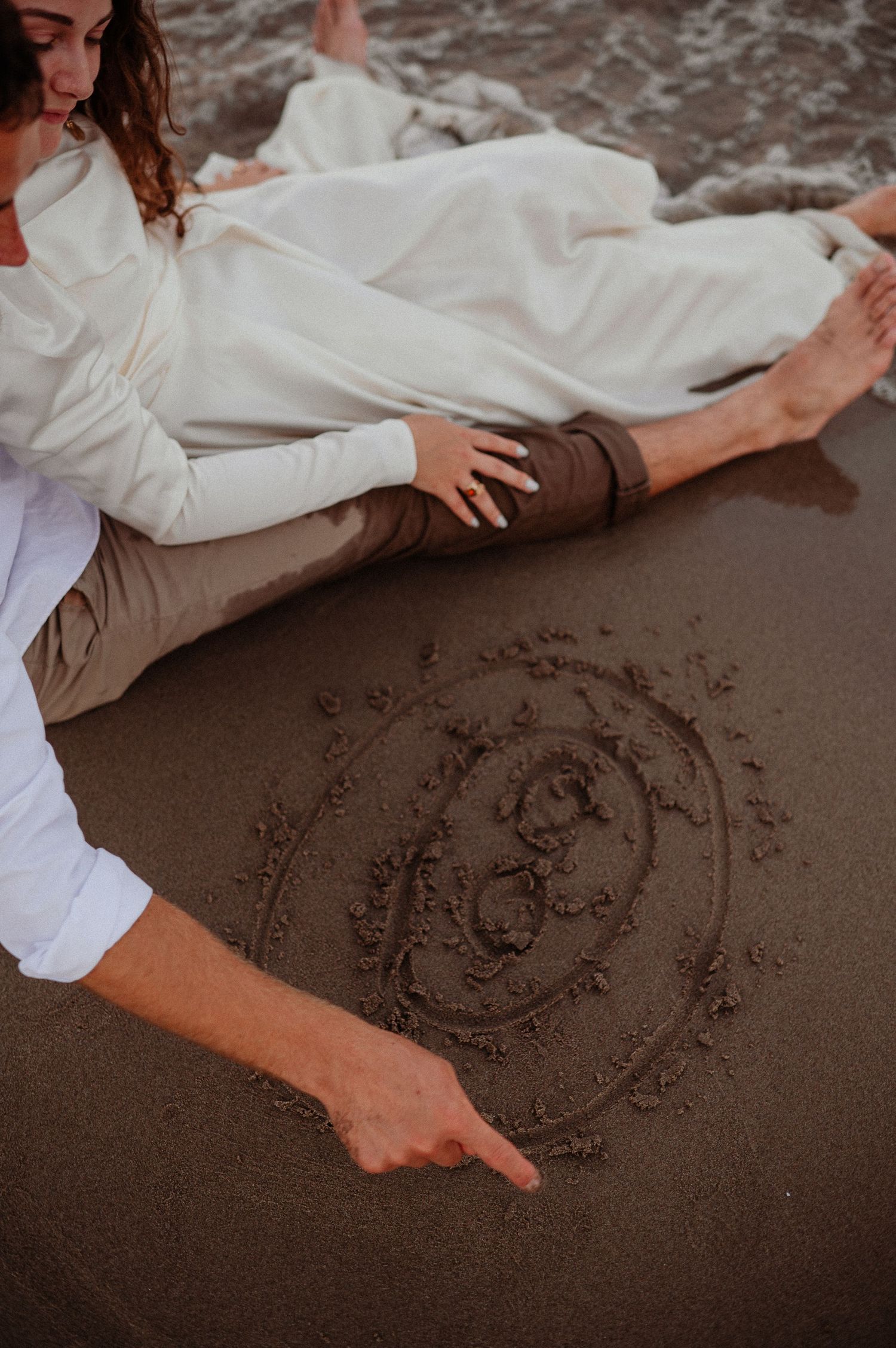 Couple sitting on beach sand pointing at a heart drawing with infinity symbol during romantic seaside photo session.