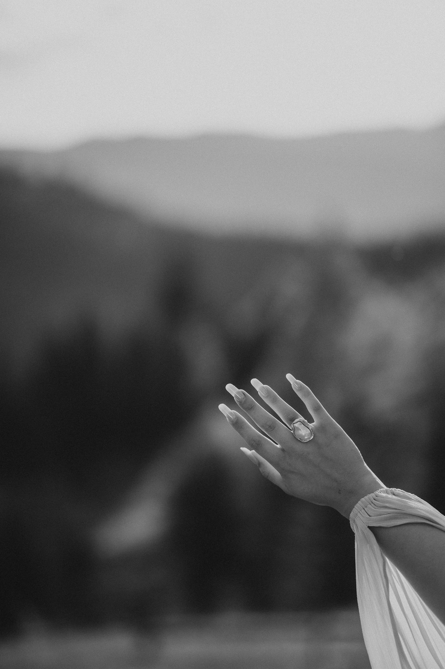 Black and white close-up of a womans hand raised showing an engagement ring with mountain backdrop.