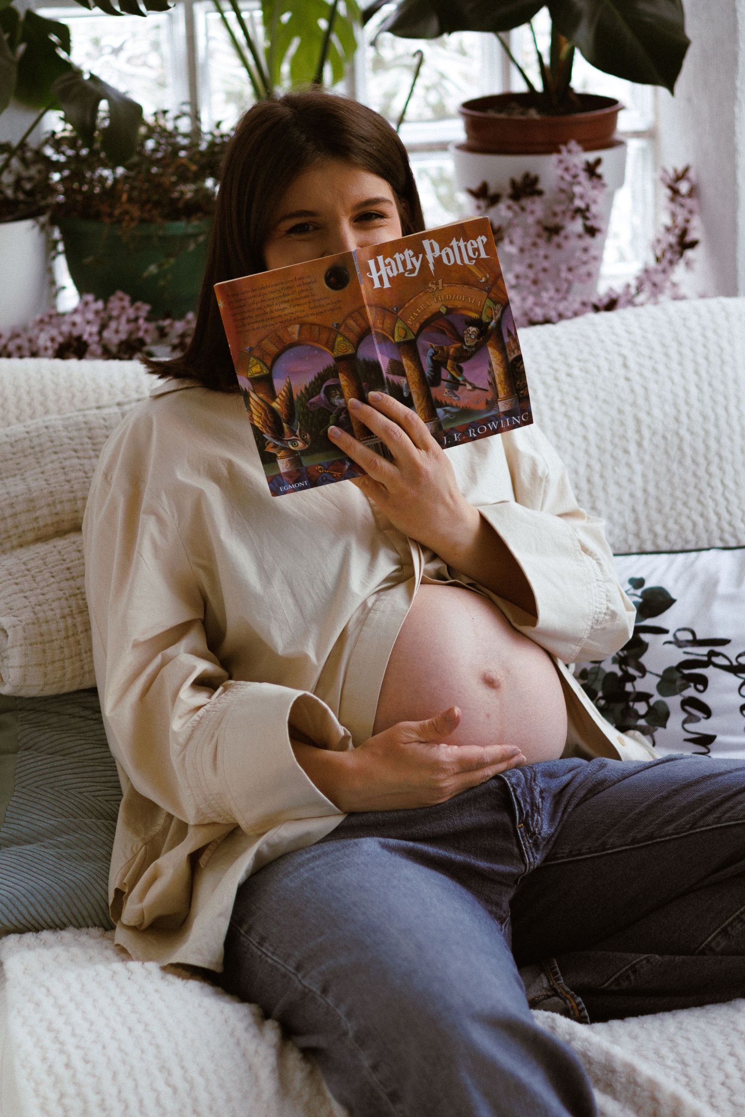 Pregnant woman reading Harry Potter book while cradling baby bump on couch indoors.