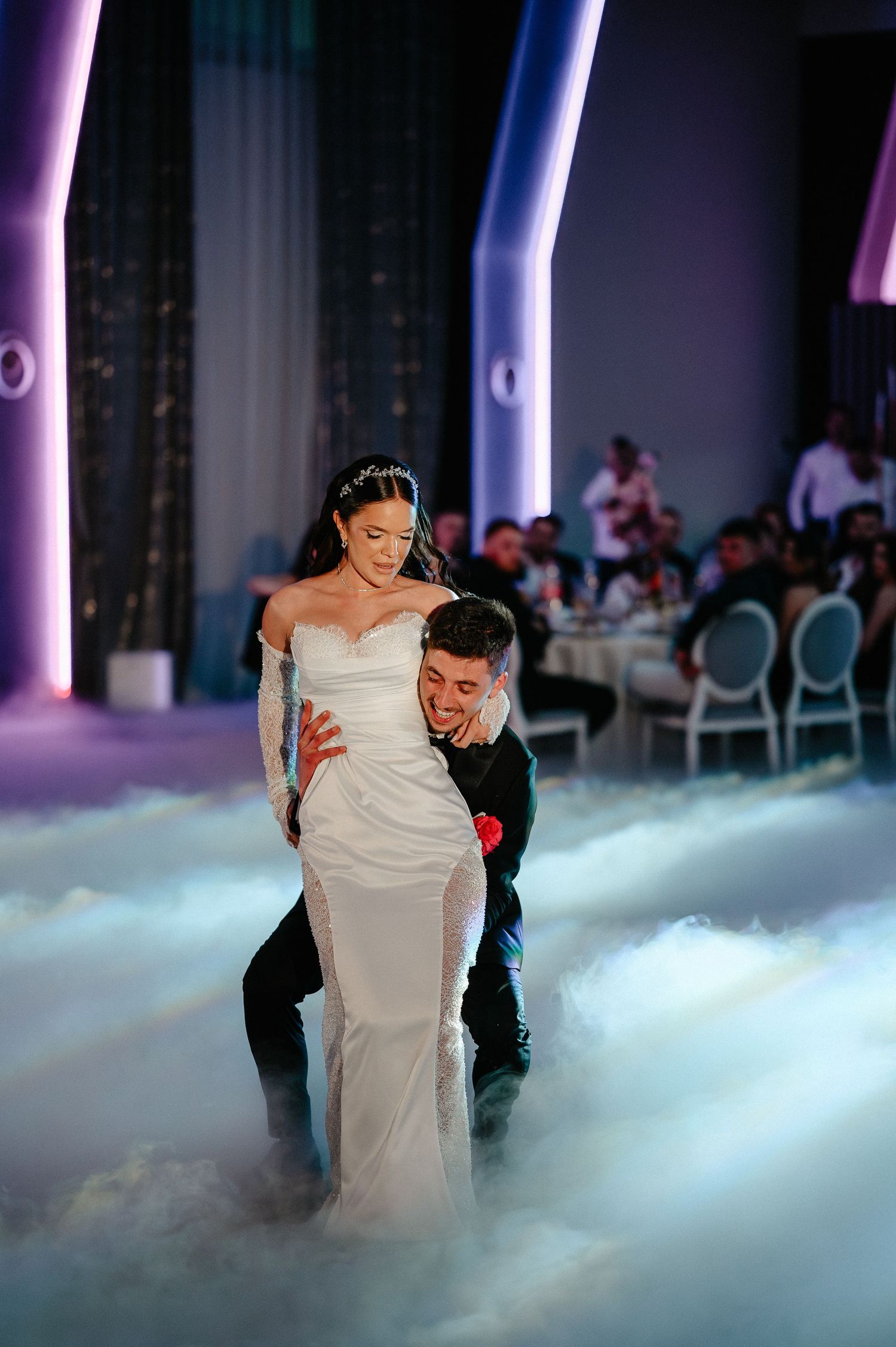 Groom lifts bride in white wedding gown during dramatic first dance surrounded by dry ice fog effects.
