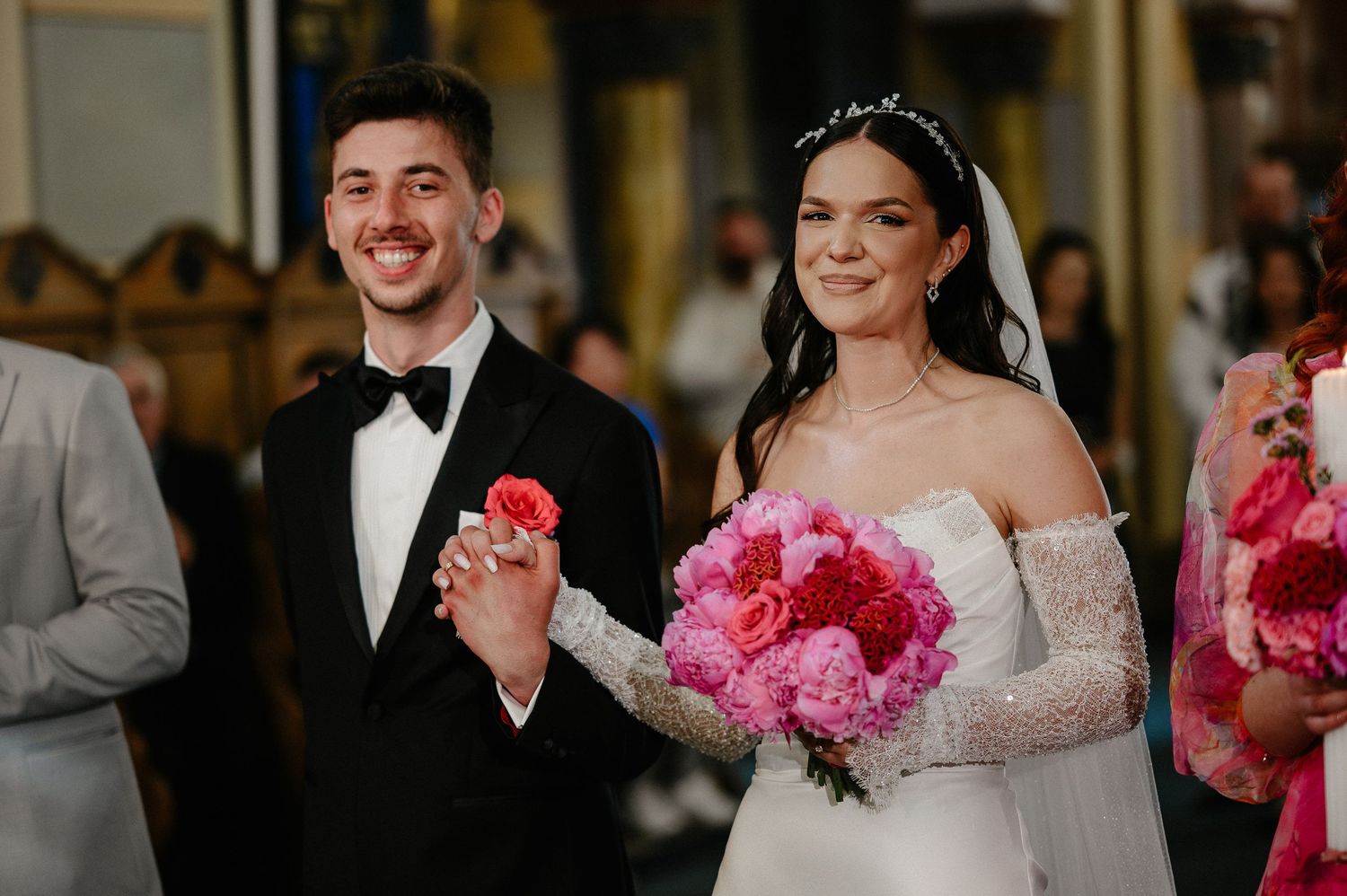 Happy bride and groom hold hands smiling after Orthodox wedding ceremony with pink floral bouquet.
