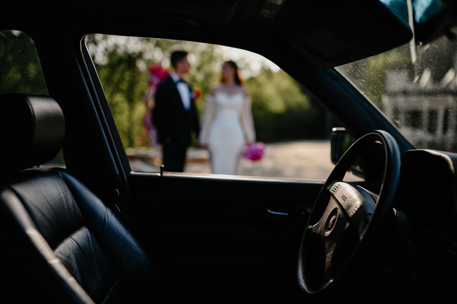 Wedding couple viewed through classic car window, bride in white gown holding pink bouquet outdoors.