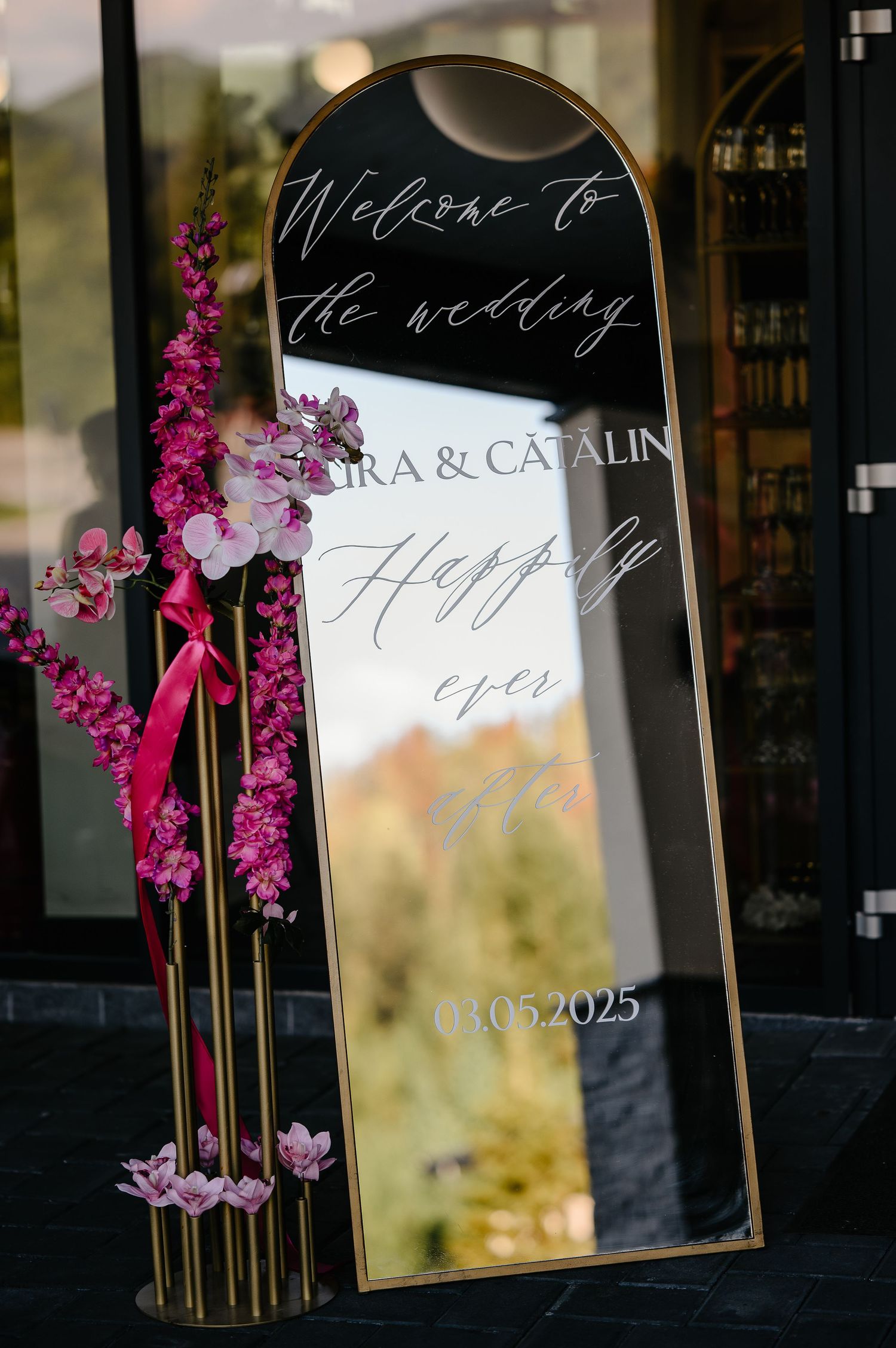 Elegant arched mirror wedding welcome sign for Ira and Catalin dated 03.05.2025 adorned with pink floral arrangements.
