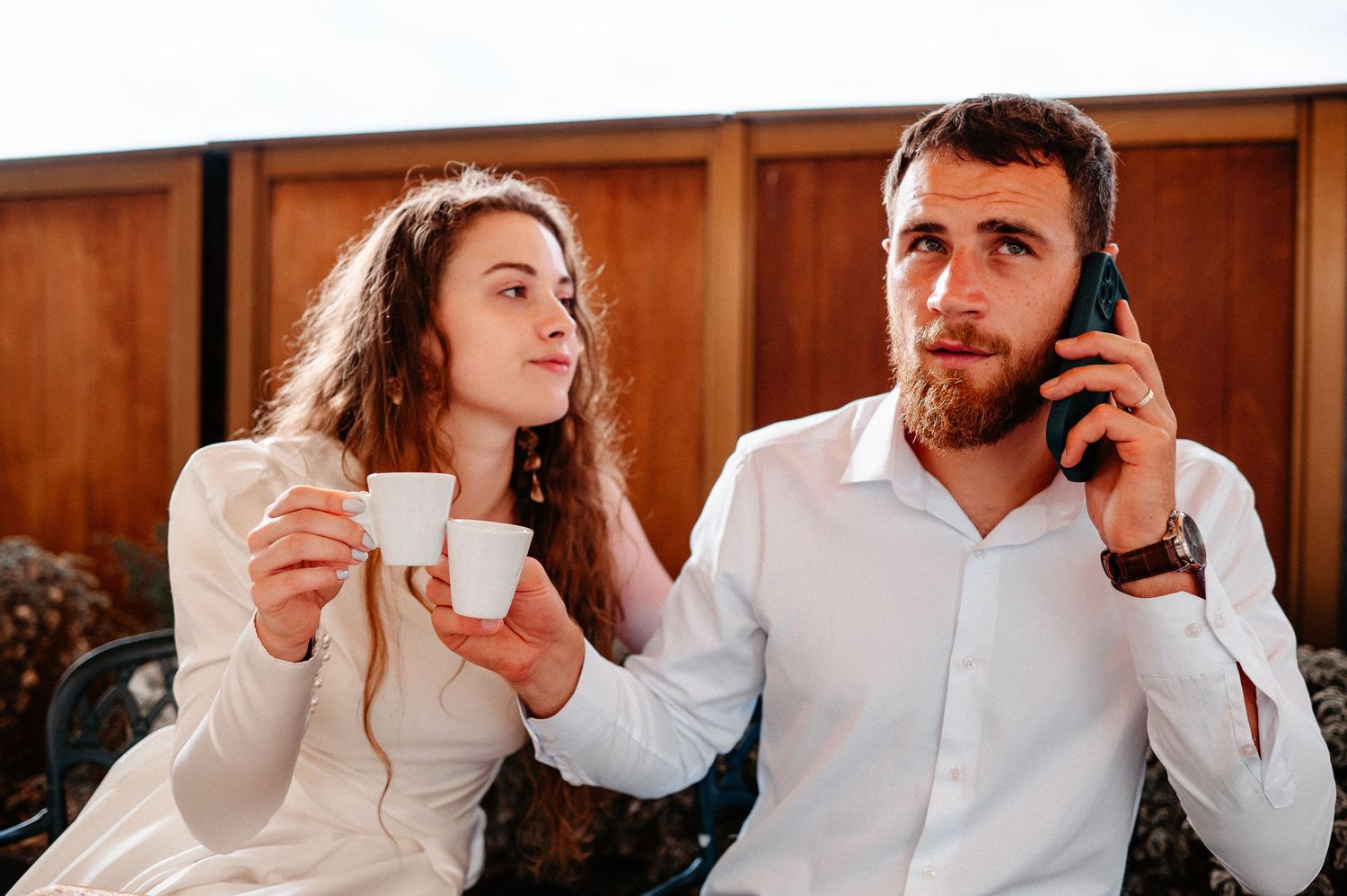 Bride and bearded groom on terrace, she holds espresso cups while he talks on phone.