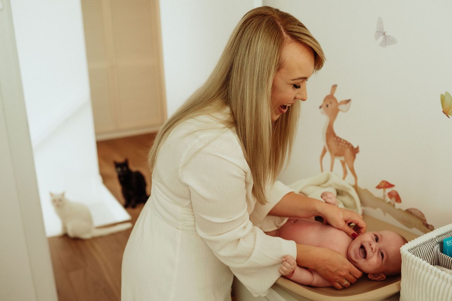 Smiling blonde mother changing newborn baby on dresser in white nursery with cat nearby.