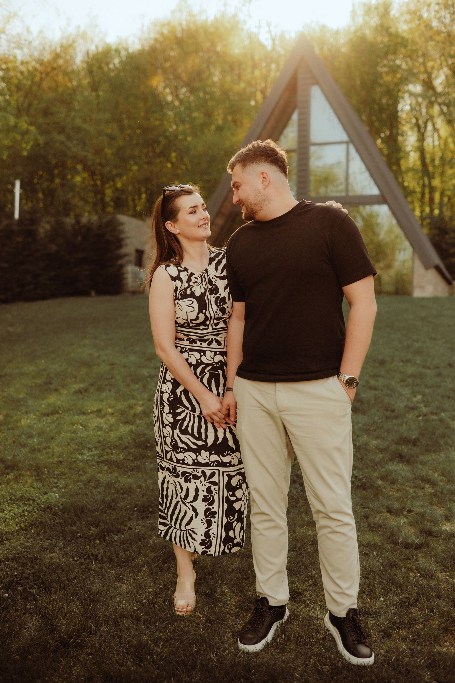 Happy couple holding hands and smiling at each other in a sunny garden near an A-frame house.