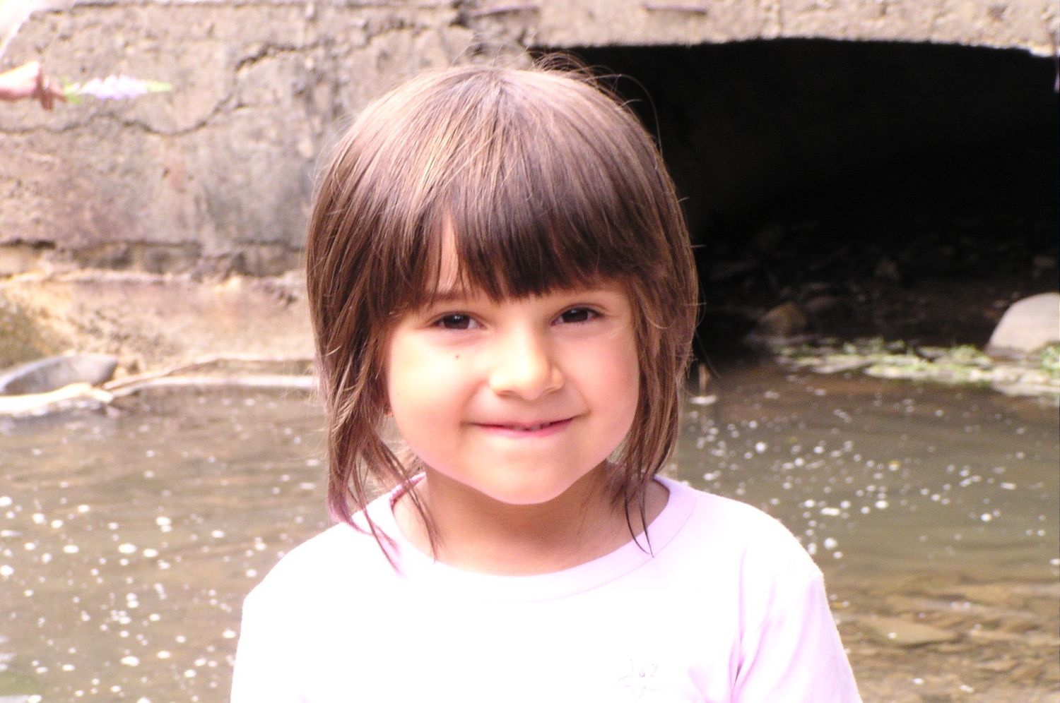 Cute smiling young girl with bangs posing outdoors near a stream on a cloudy day.