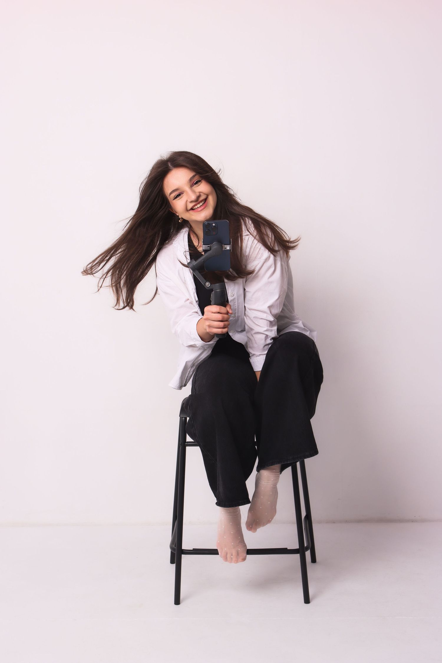 A laughing woman with flowing hair sits on stool holding camera stabilizer on white background.