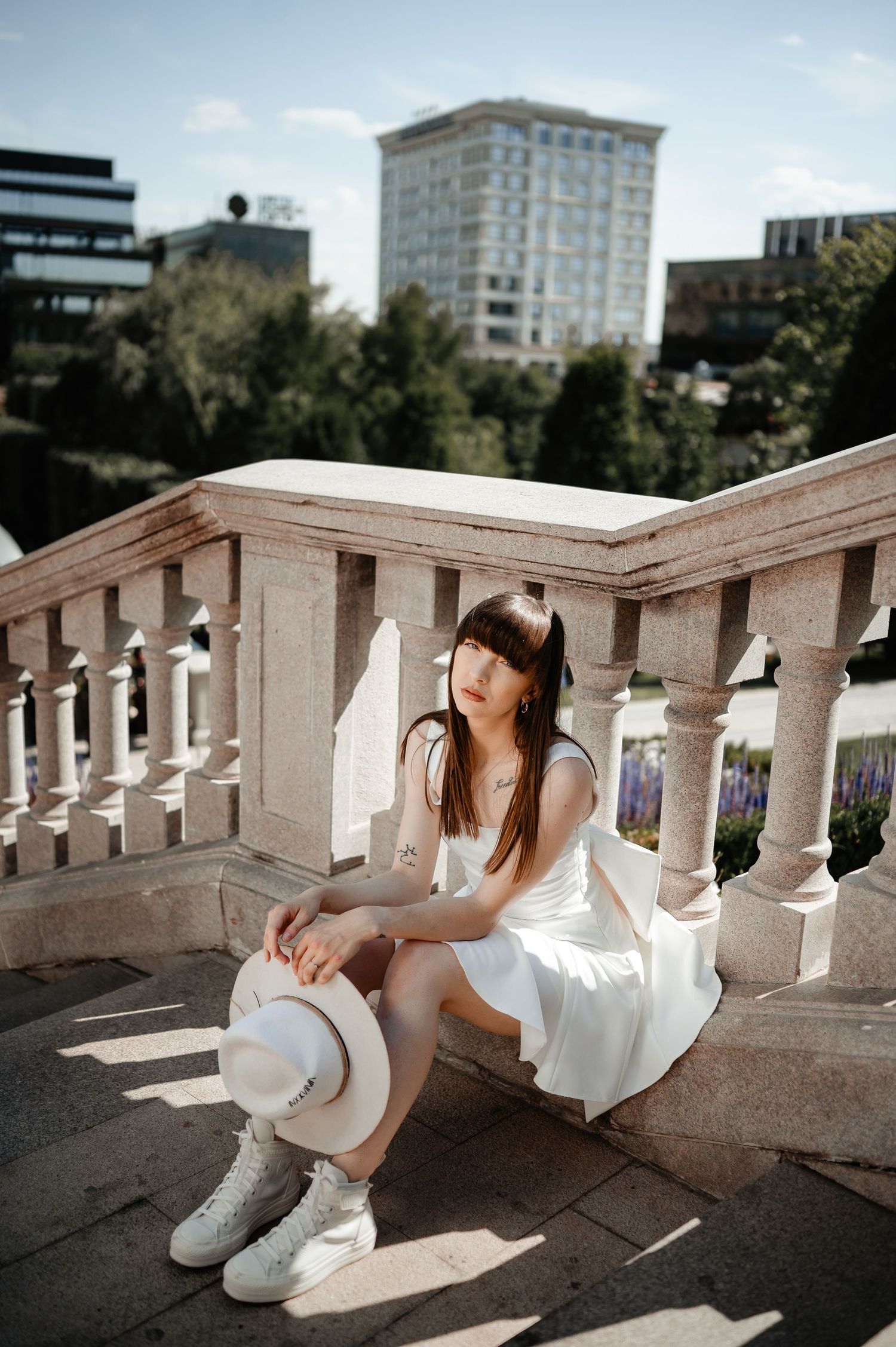 Young woman in white dress and sneakers sits on a stone balustrade with urban buildings in the background.