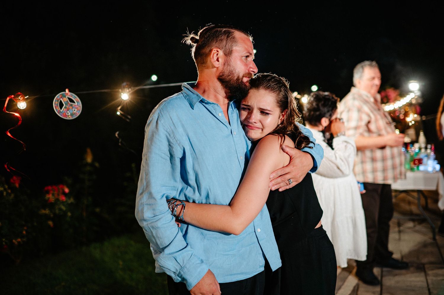 A couple shares a tender embrace at an outdoor evening celebration with string lights.