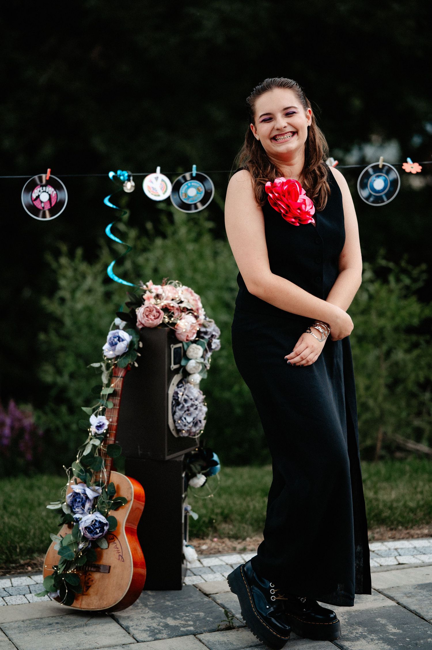 Smiling young woman in black maxi dress with rose corsage posing beside decorated guitar at party.