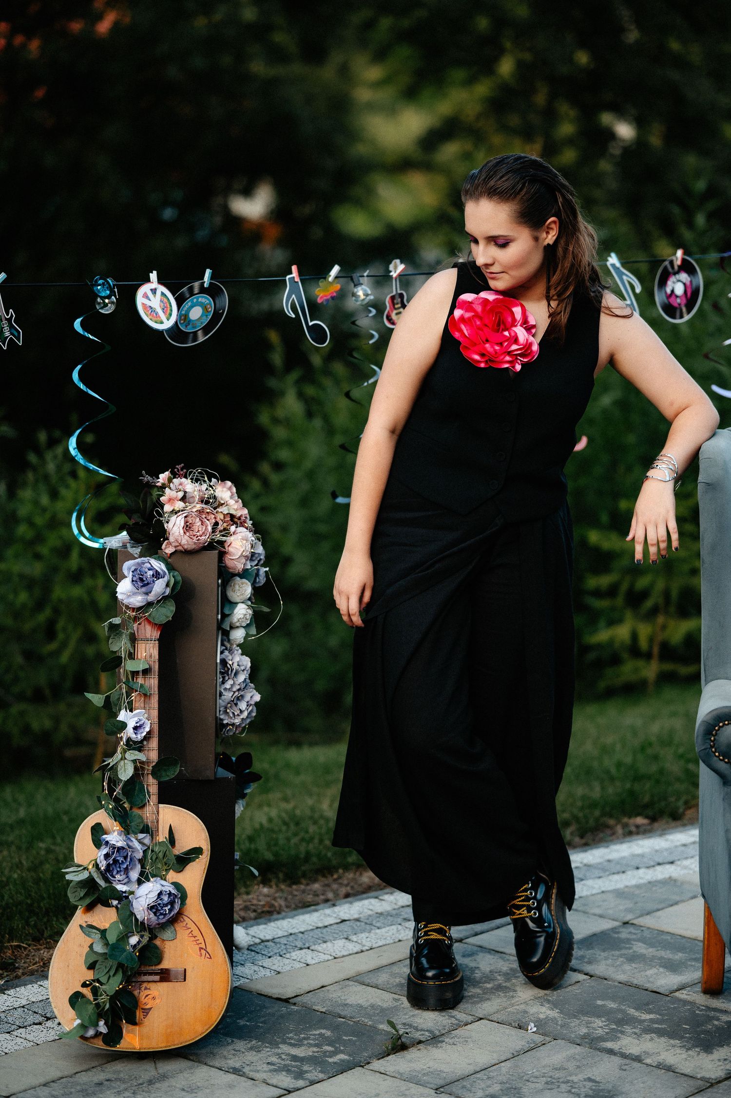 Young woman in black maxi dress with red flower walking beside a floral-decorated guitar outdoors.