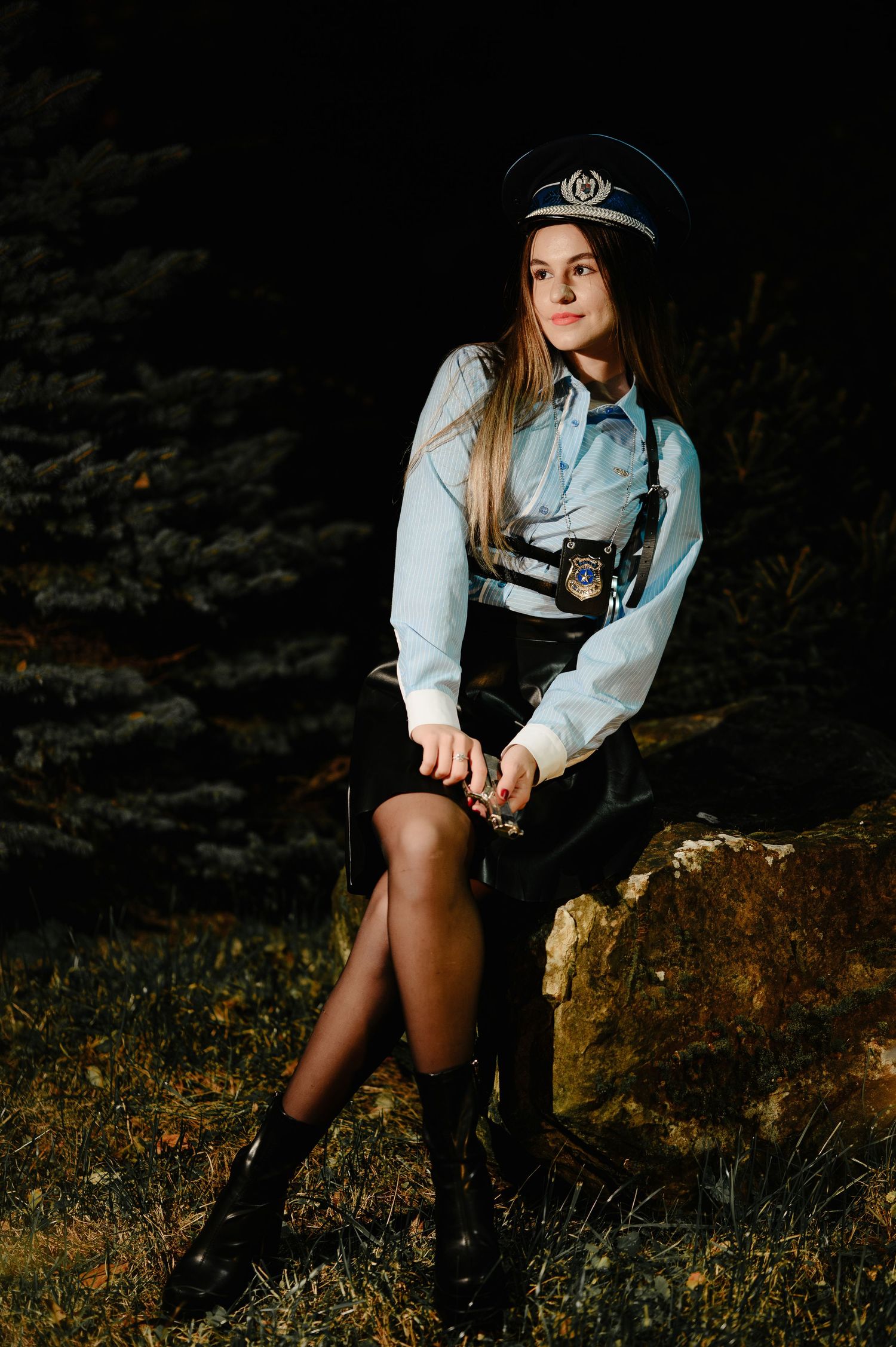Young woman in police officer costume with cap and badge poses on rock at night.