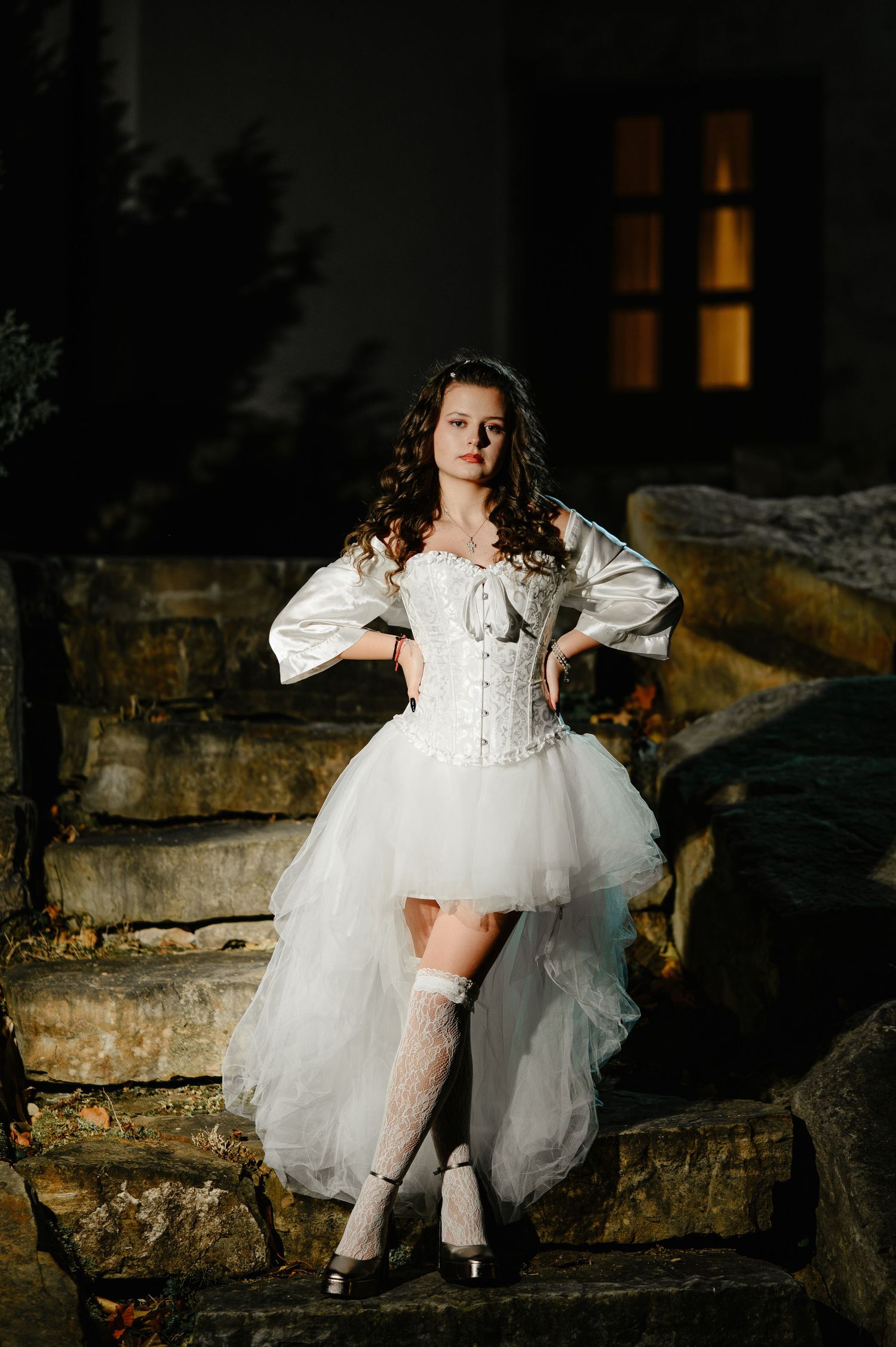 Young woman in elegant white corset Halloween bride costume posing confidently on stone steps at night.