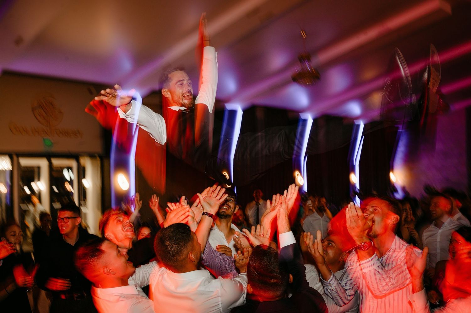 Energetic wedding guest lifted by crowd during a lively reception celebration with colorful party lights.