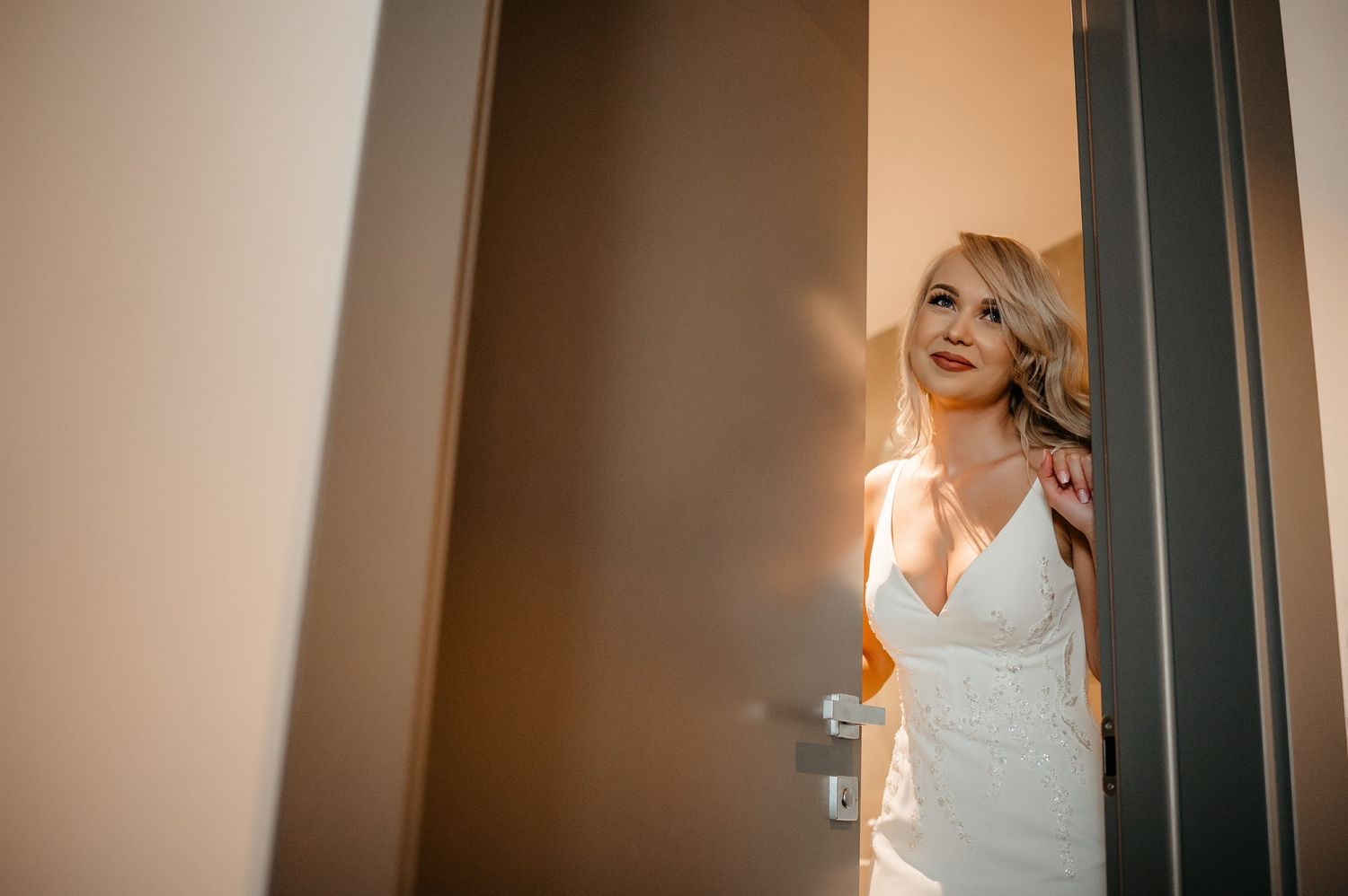 Radiant blonde bride in elegant white wedding dress standing in golden sunlit doorway.