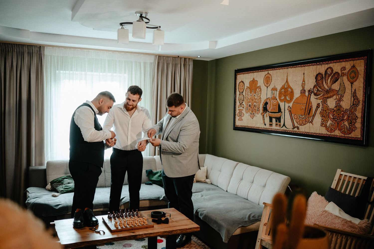 Three men in formal attire helping each other get dressed in a hotel room before a wedding.