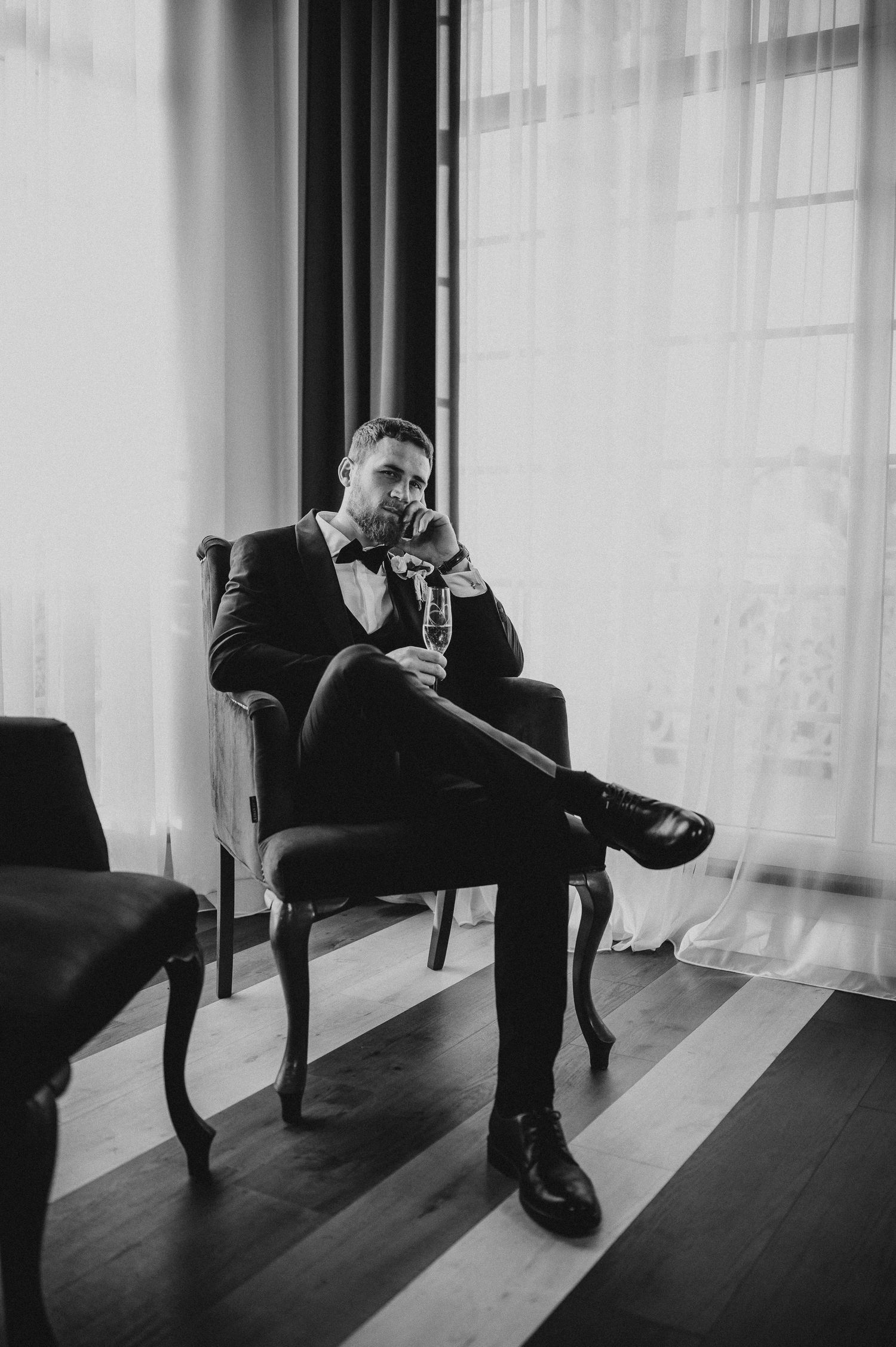 Black and white portrait of groom in tuxedo sitting in armchair holding champagne glass by window.