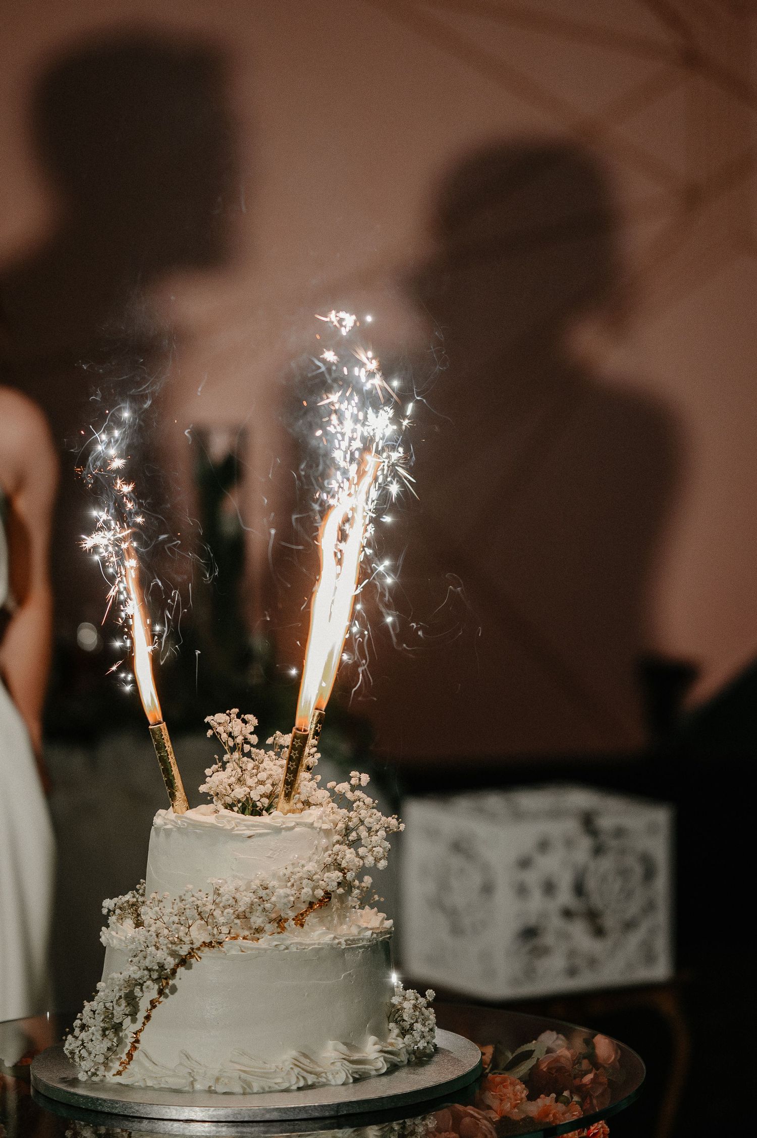 Elegant two-tier white wedding cake decorated with baby breath flowers and glowing sparklers on top.