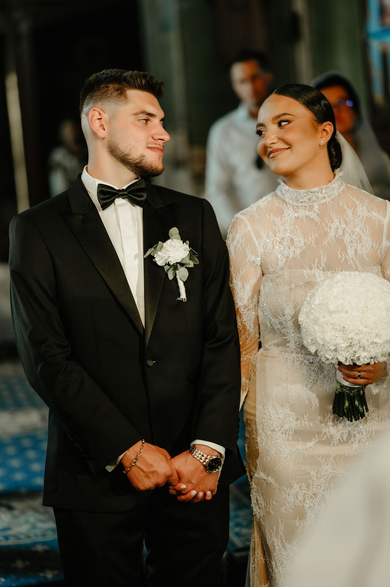 Groom in black tuxedo and bride in lace gown smile together during church wedding ceremony.