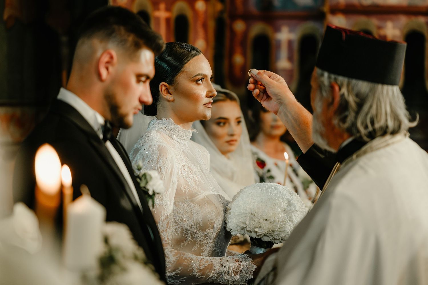 Bride and groom receiving Orthodox wedding ceremony blessing from priest with candles.