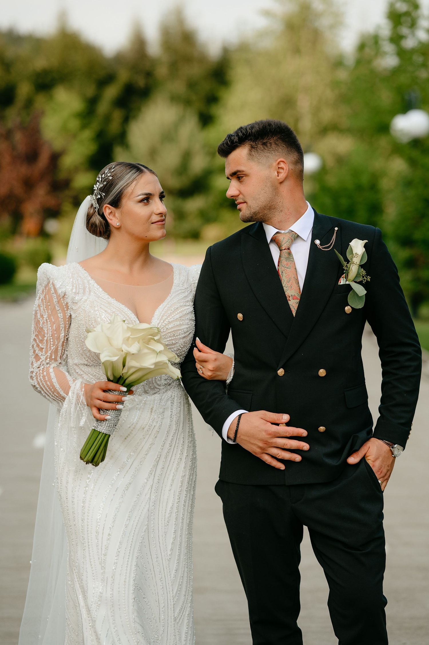 Bride in beaded gown holding white calla lily bouquet walks arm-in-arm with groom in black double-breasted suit.