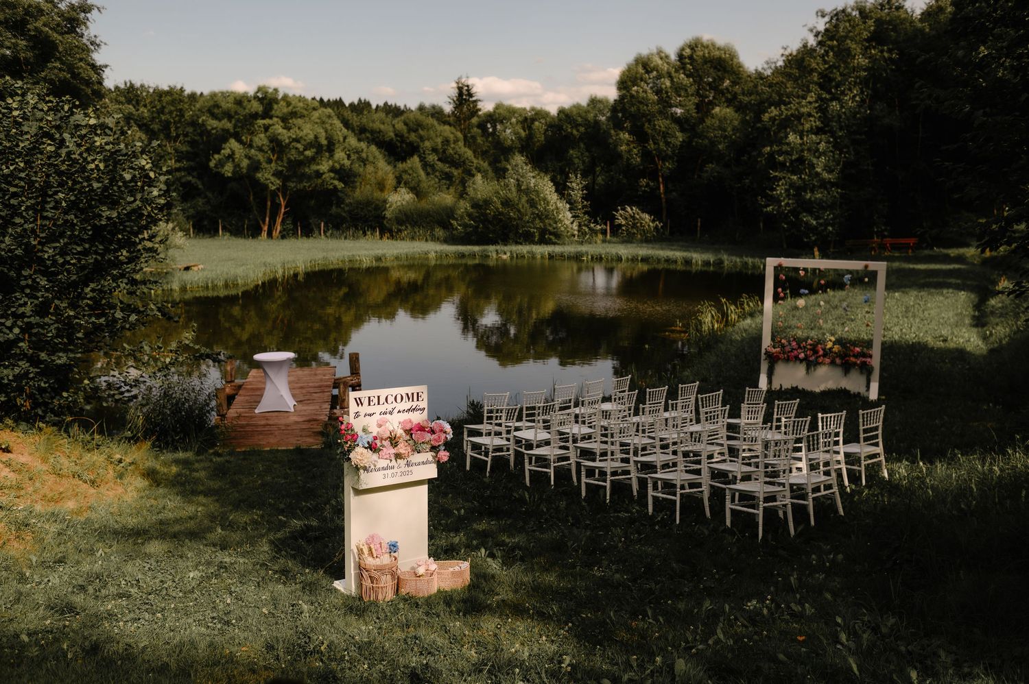 Outdoor wedding ceremony setup with white chairs and floral arch beside a calm reflective pond.