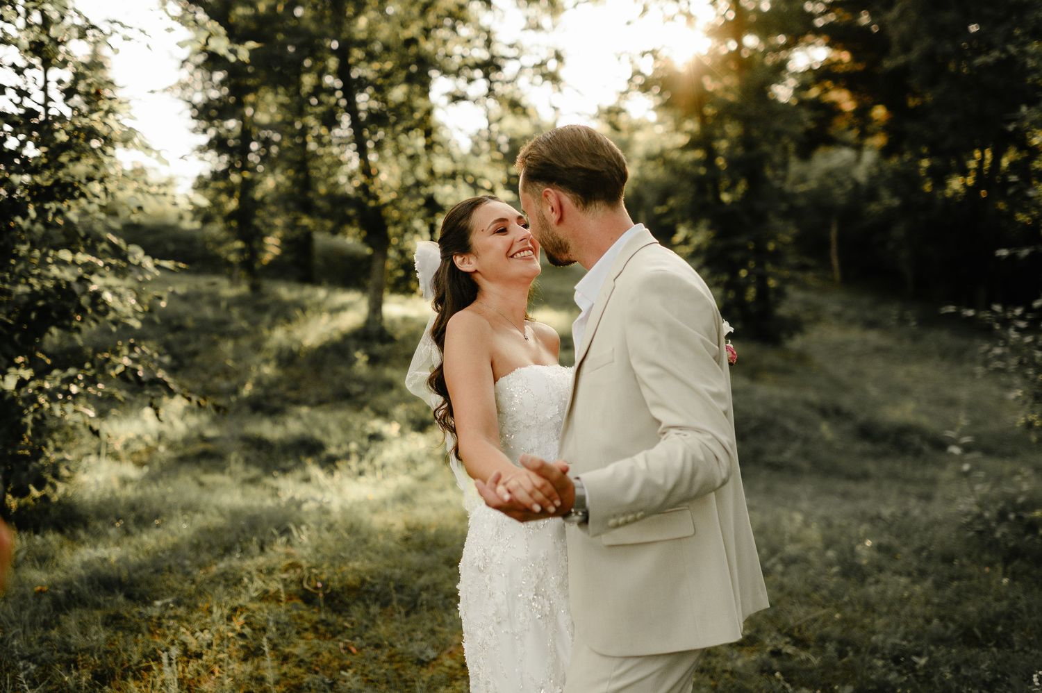 Groom kissing smiling bride on cheek in golden sunlit forest during romantic outdoor wedding portrait.