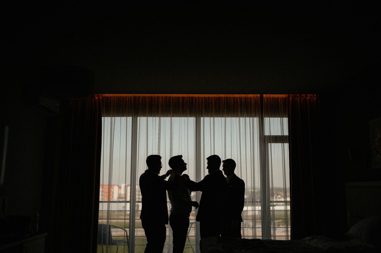 Silhouette of four groomsmen gathered together by curtained window during wedding morning preparations.