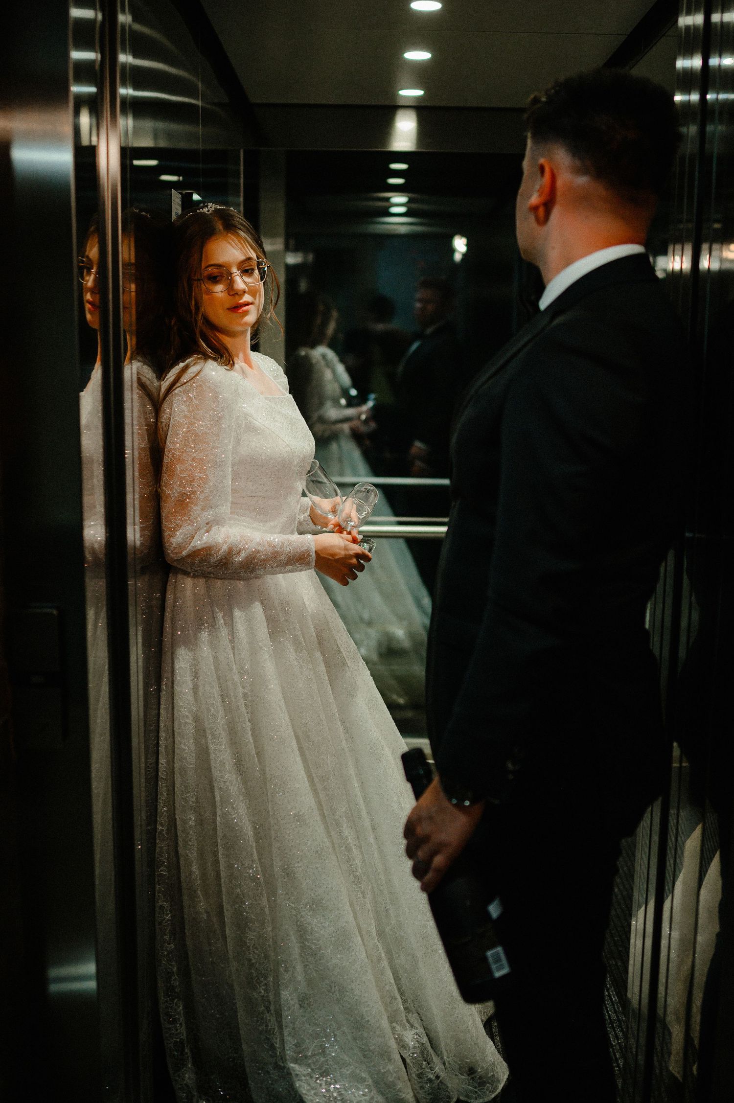 Bride in a long-sleeve lace wedding dress stands in an elevator holding champagne glasses.