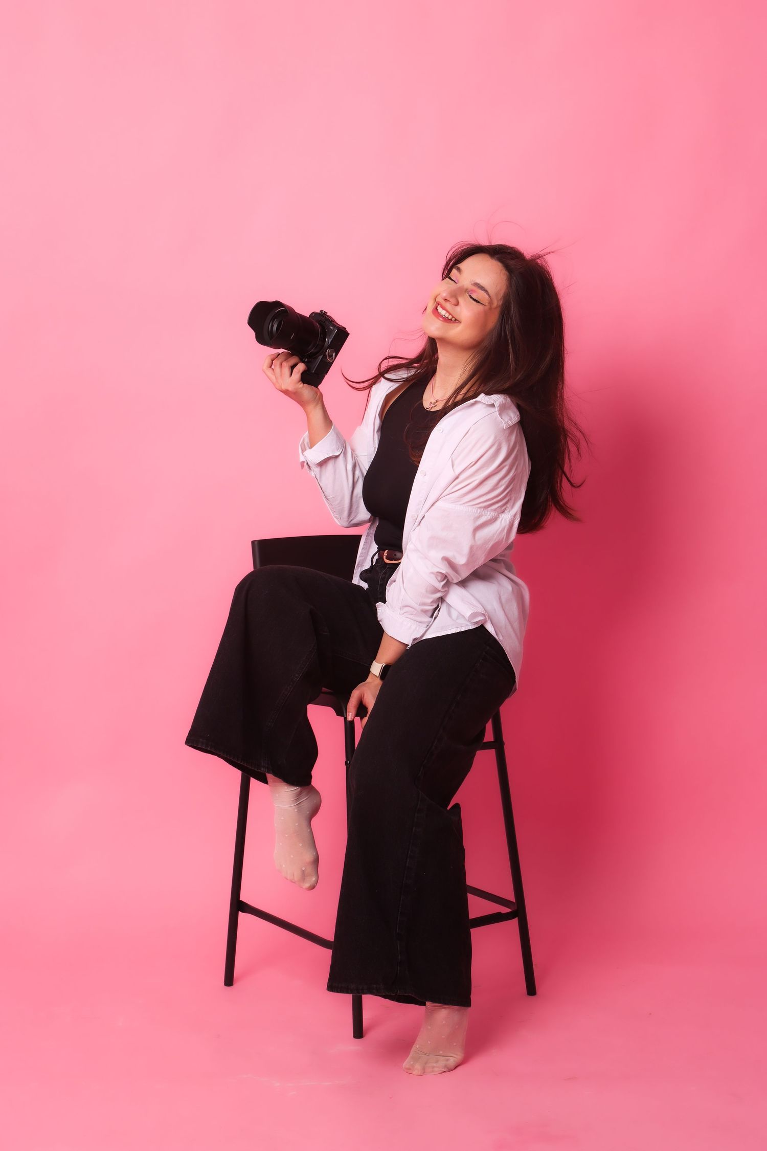Young woman photographer sitting on stool holding camera, wearing white shirt and black pants against pink background.