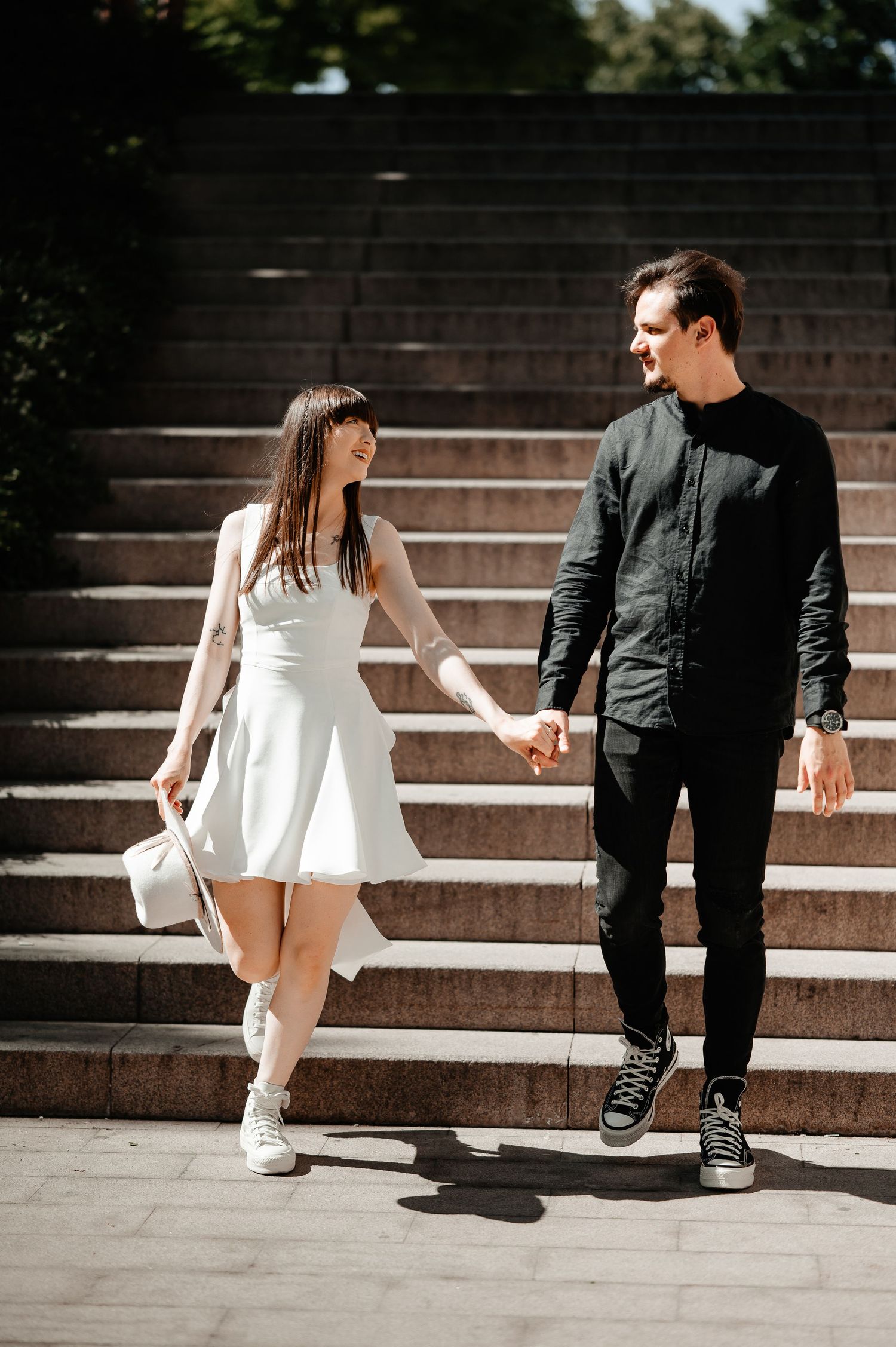Smiling couple holding hands and walking down sunlit stone steps, woman in white dress carrying a hat.