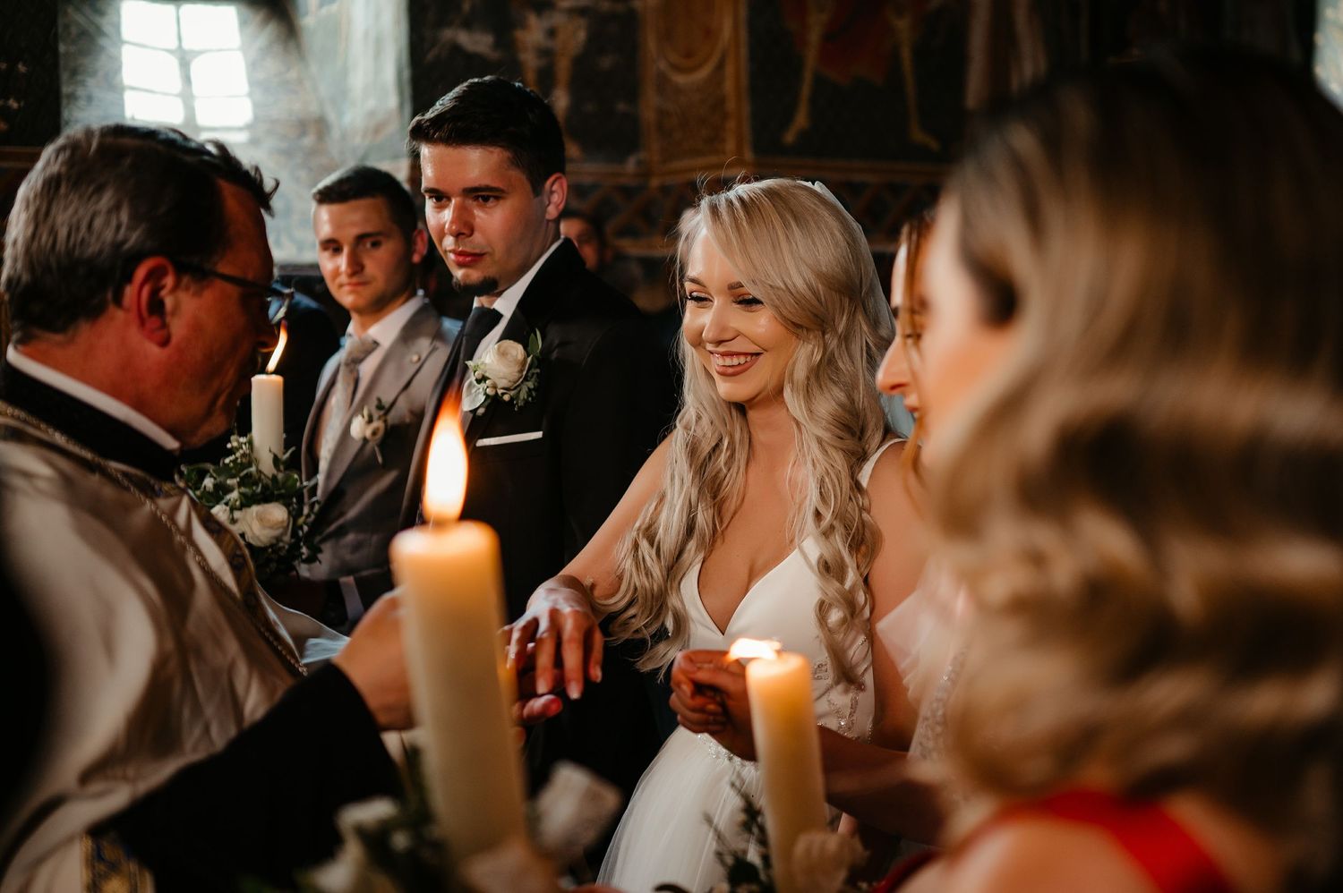 Smiling bride in white gown seated with groom during candlelit Orthodox wedding ceremony indoors.