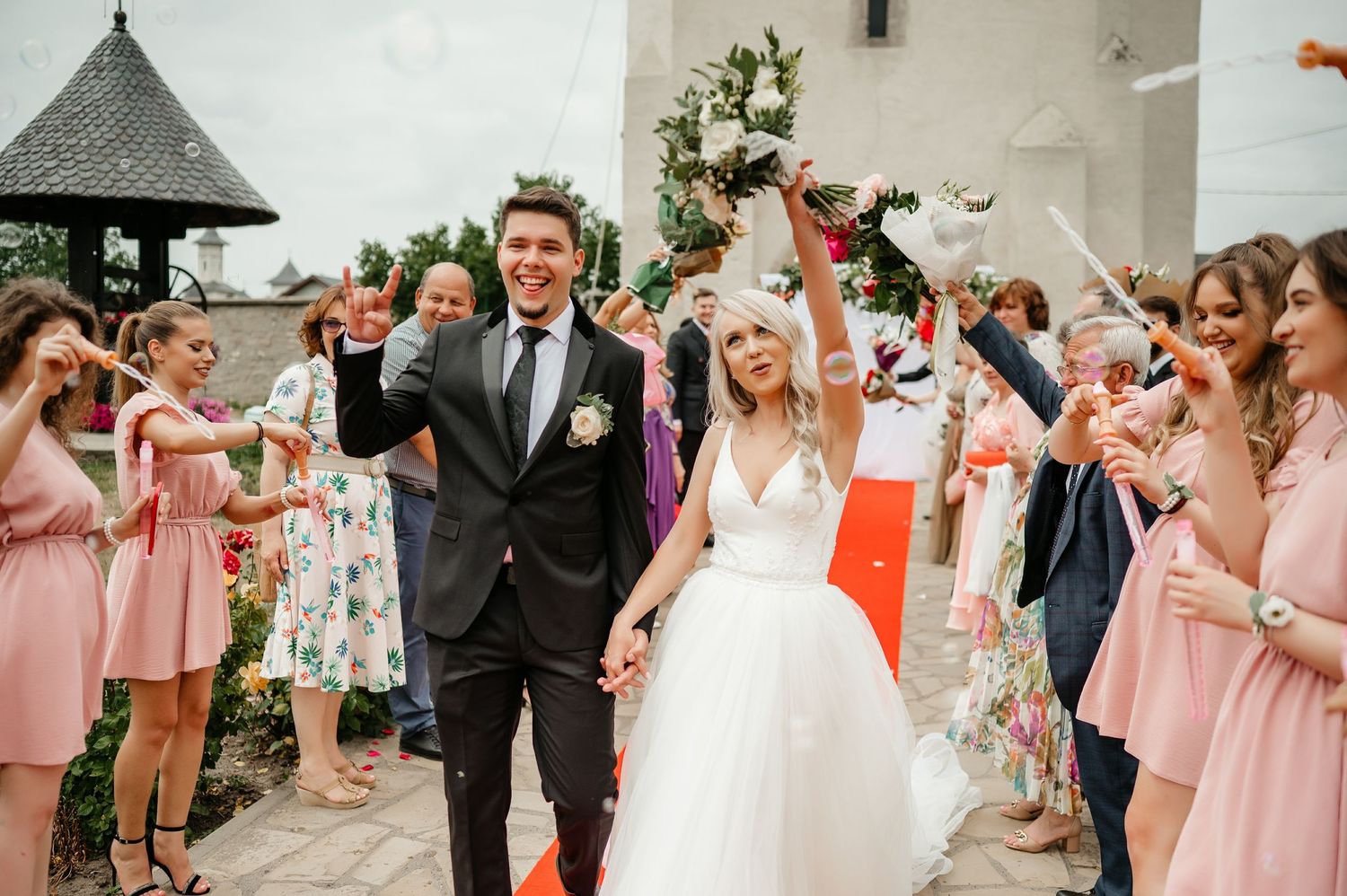 Joyful bride and groom raise their hands in celebration as bridesmaids in pink dresses cheer.