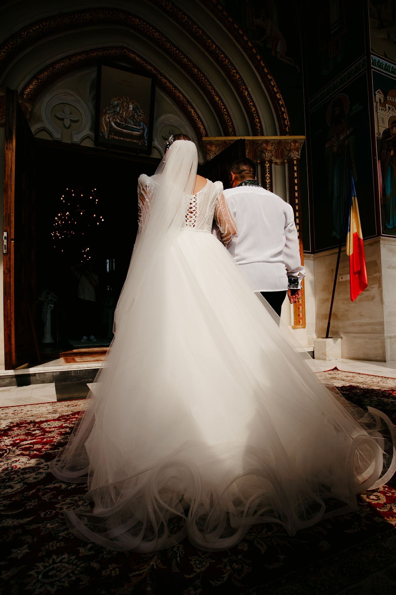 Bride in full tulle wedding gown with long veil entering ornate Orthodox church with escort.