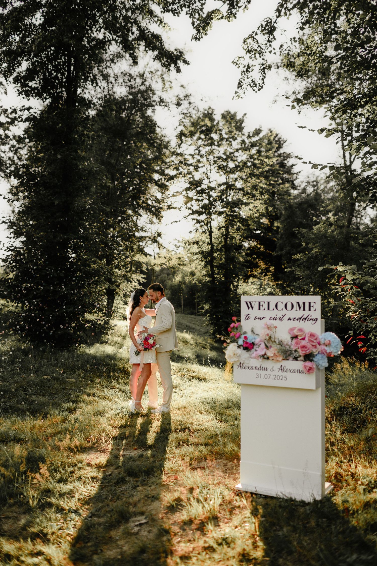 Newlywed couple embracing near welcome sign at outdoor elopement wedding ceremony in sunlit forest.